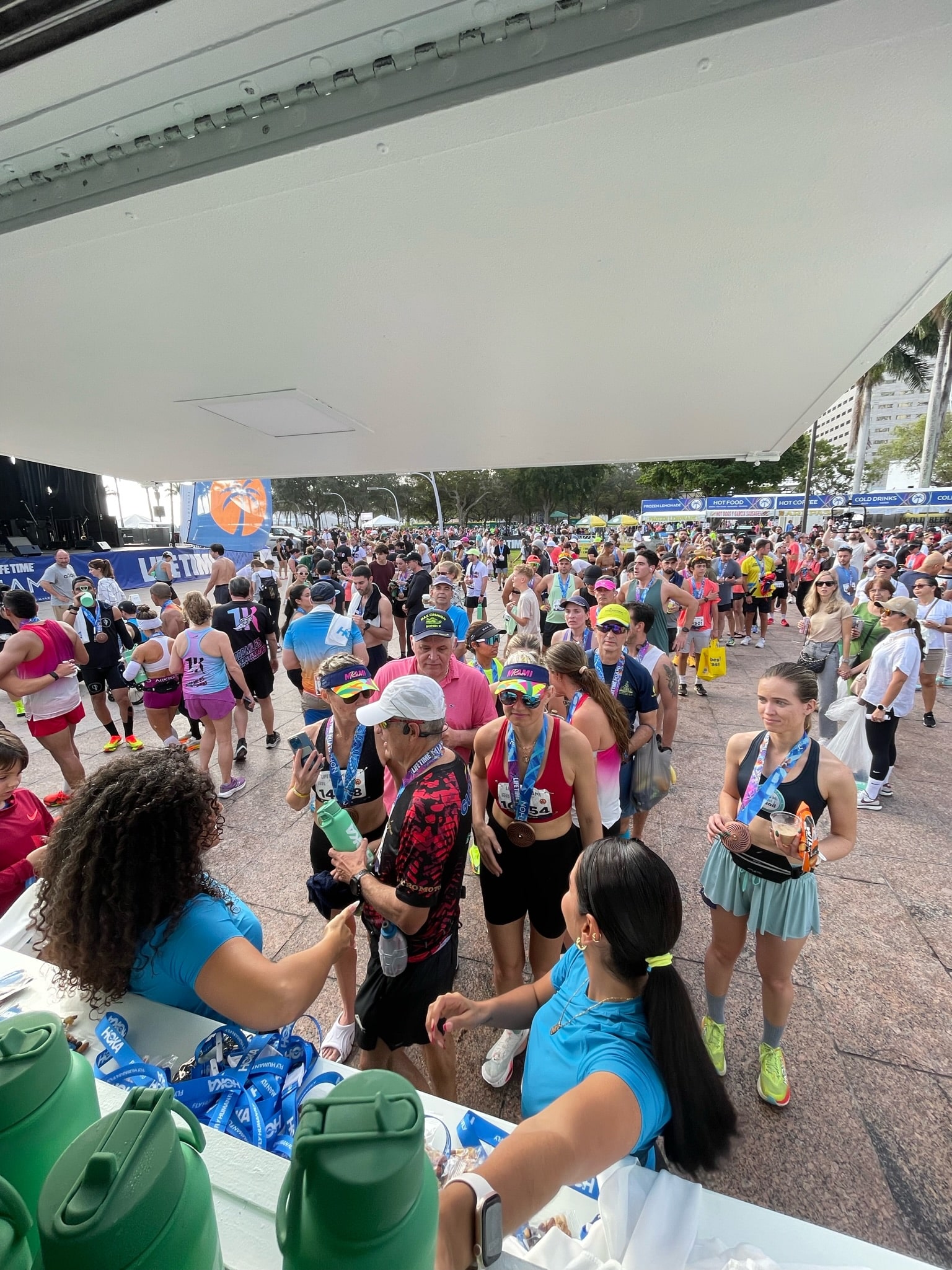 Runners with medals gather at an outdoor water station as volunteers in blue shirts serve drinks.