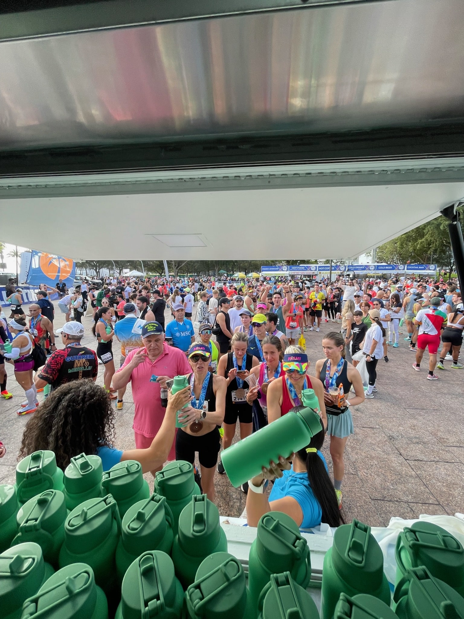 Marathon runners with medals gather at a refreshment station as volunteers give out green bottles.
