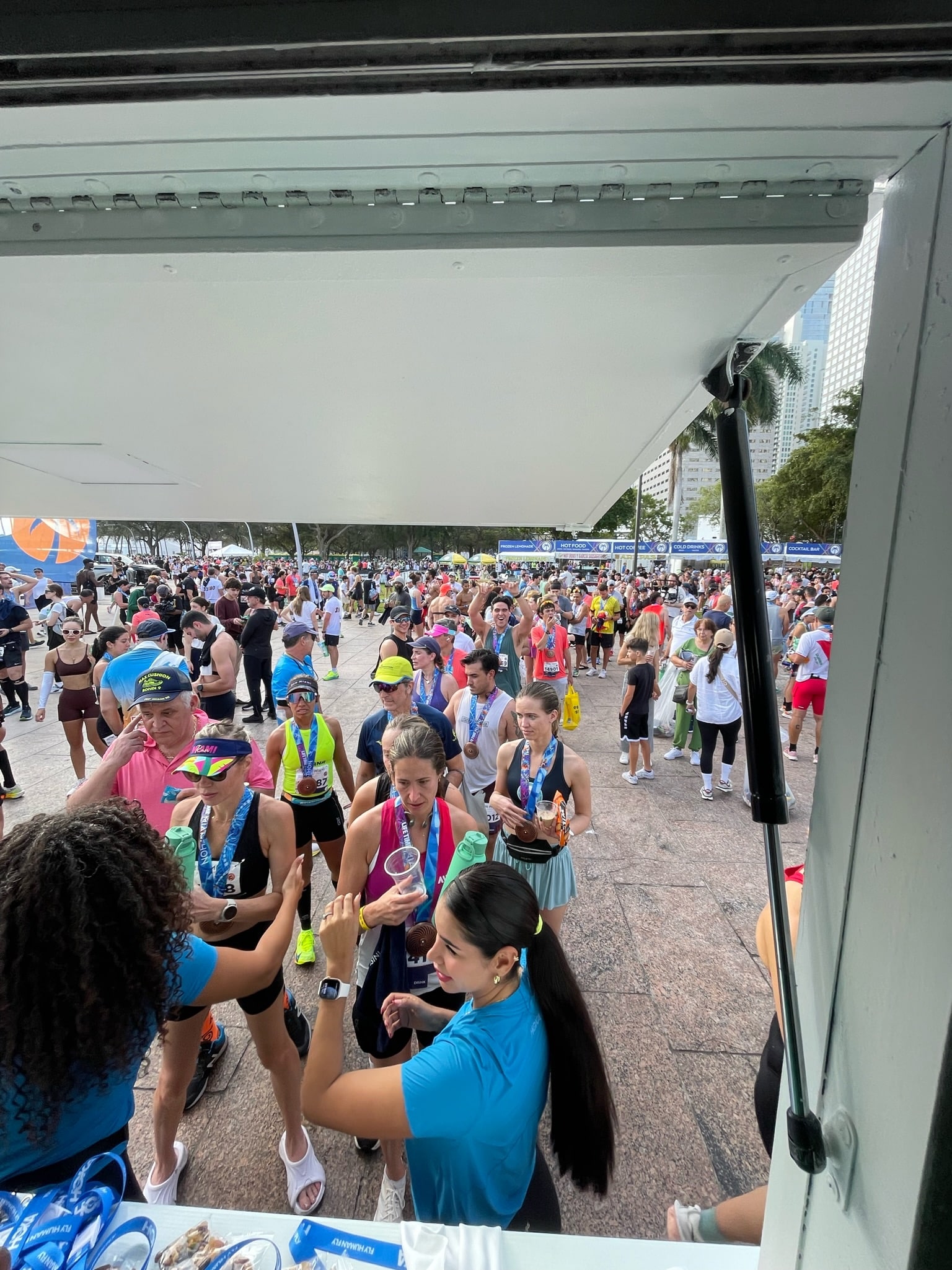 Marathon runners with medals gather at a refreshment stand as volunteers give out drinks.
