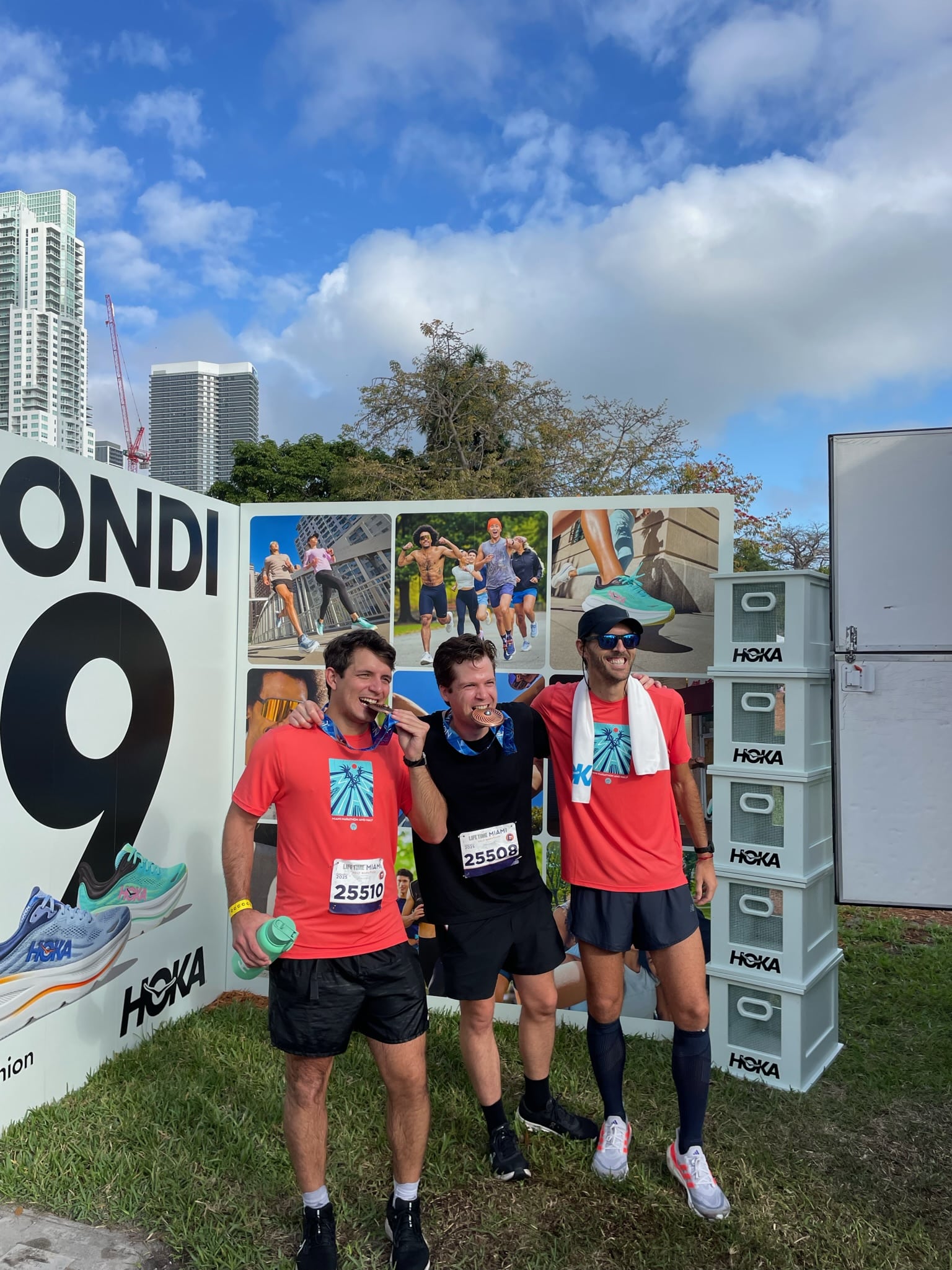 Three runners in race bibs, two in red shirts and one in black, pose by a HOKA event backdrop.