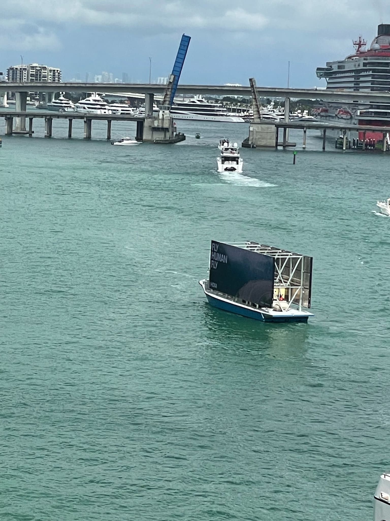 Boat with large billboard sails by a drawbridge; other boats are docked in the background.