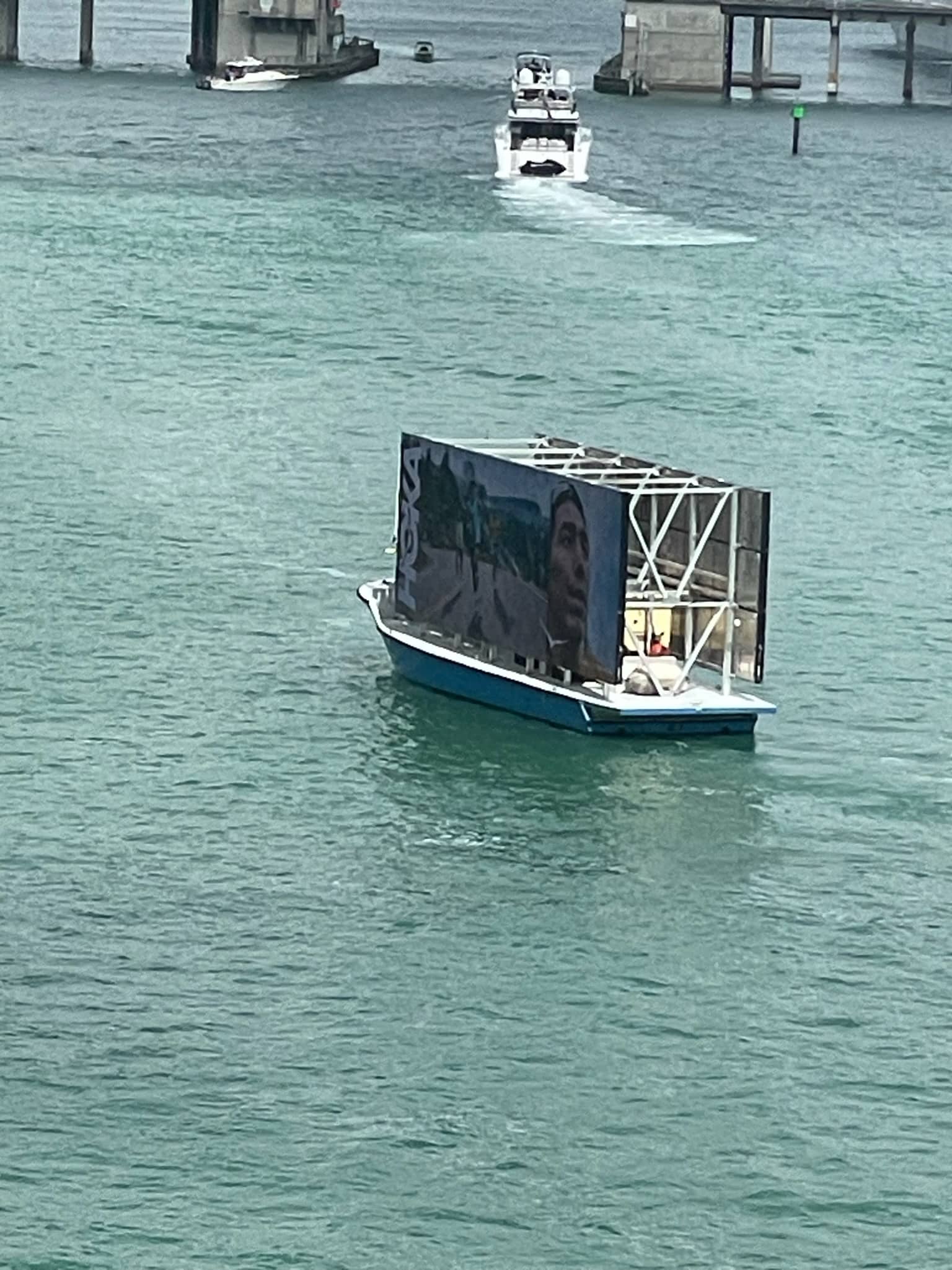 Boat with a billboard showing a face and beach scene sails on turquoise water; bridge in background.