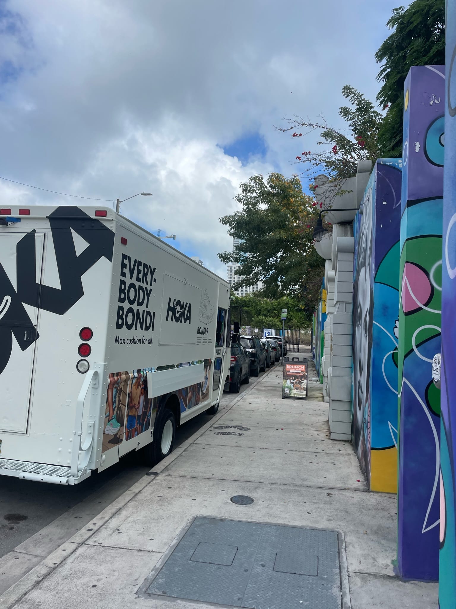 White HOKA truck with "EVERYBODY BONDI" parked by colorful murals and lined cars on city street.