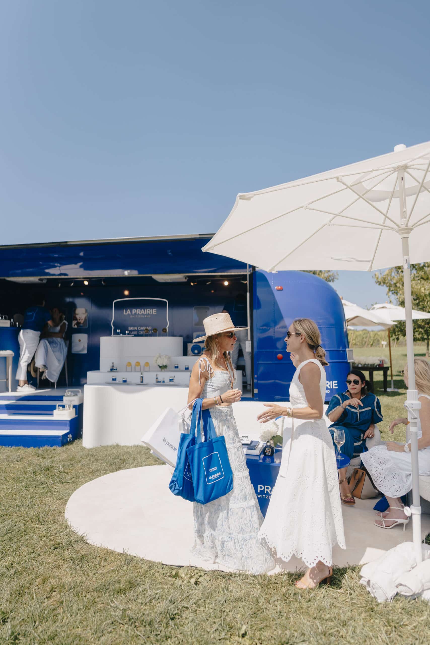 Two women in summer dresses talk outside a blue La Prairie booth; people sit under a white umbrella.