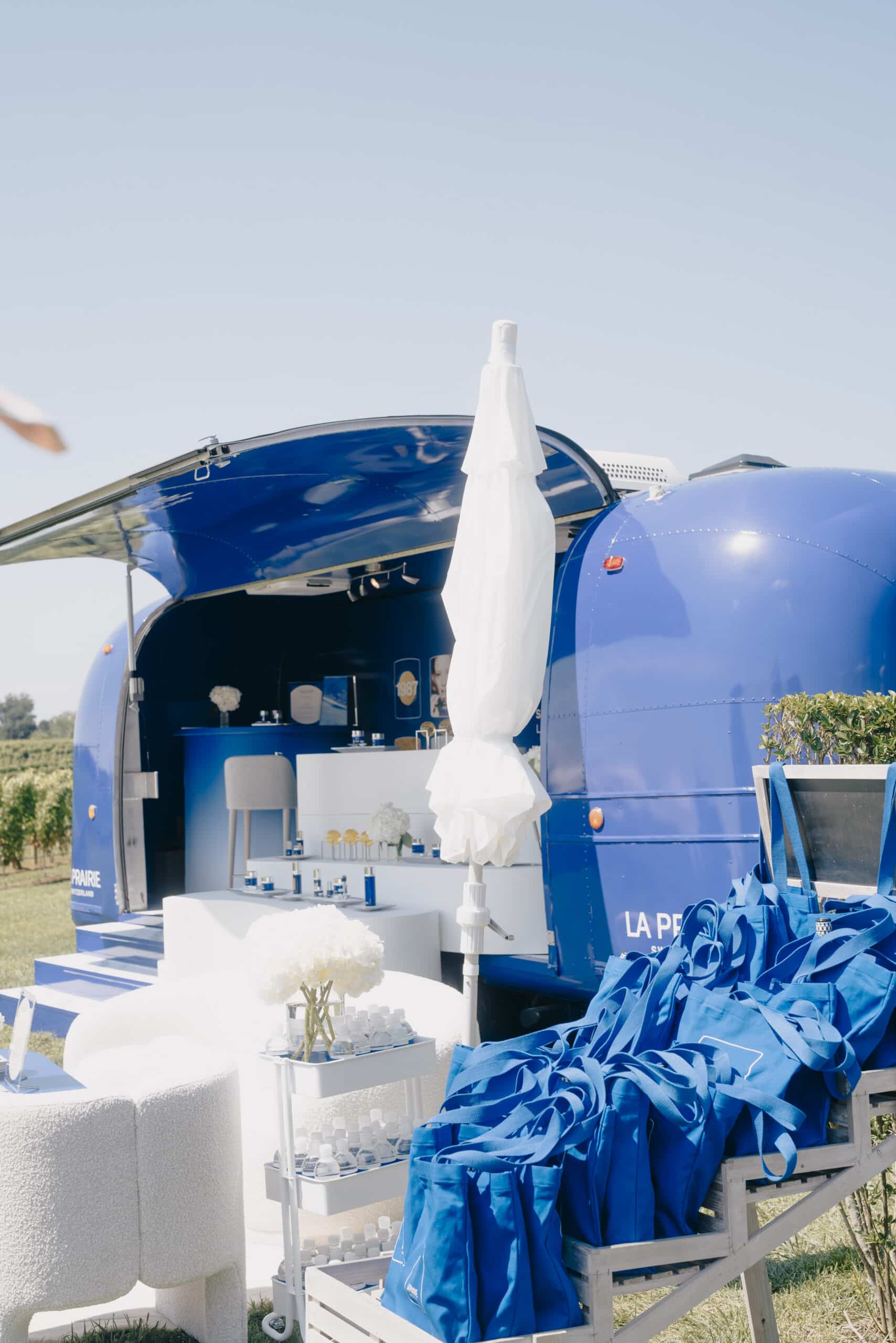 Blue Airstream trailer with open window displays skincare; umbrella and shelves on grass.