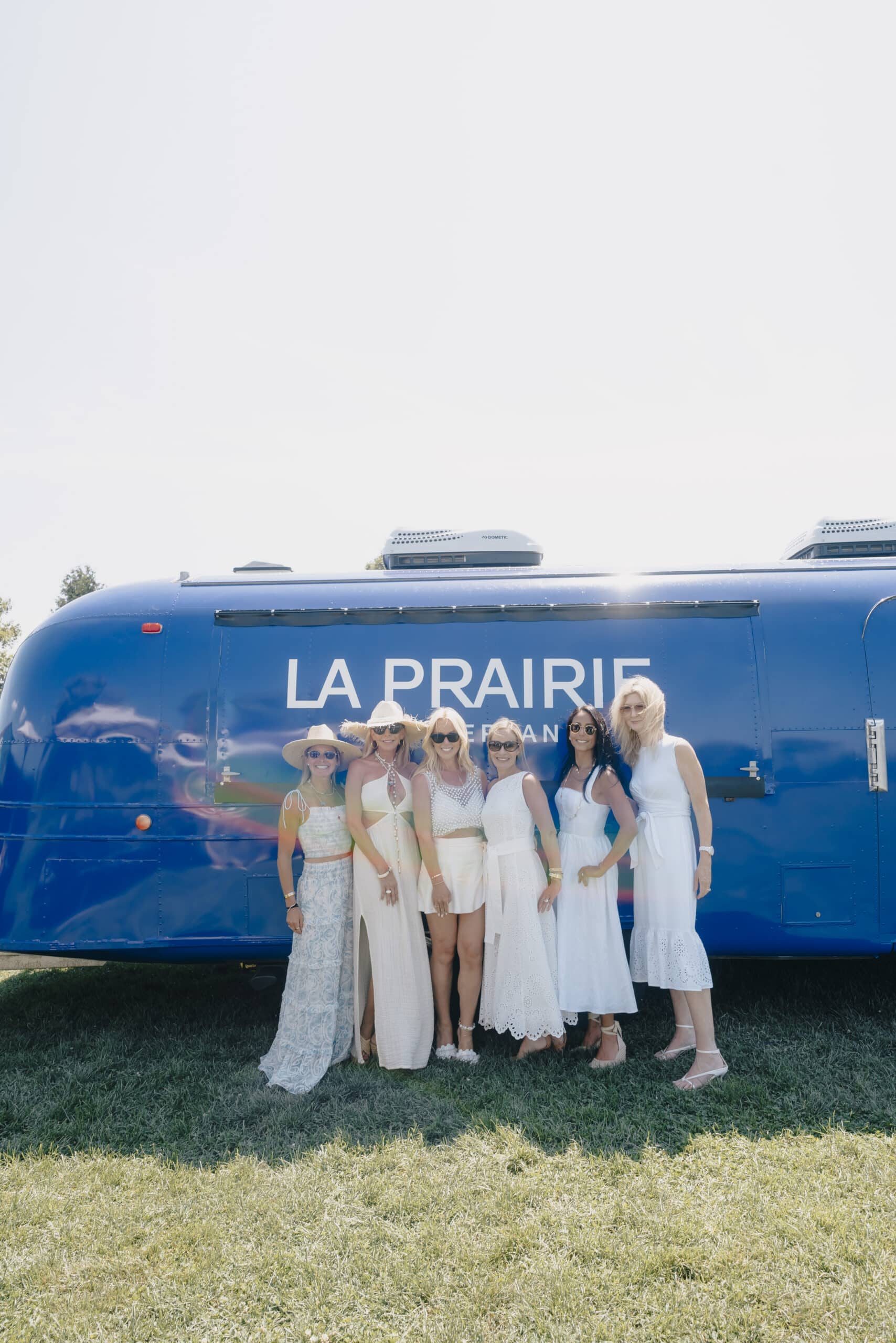 Six women in summer dresses smile together outside a large blue Airstream labeled La Prairie.