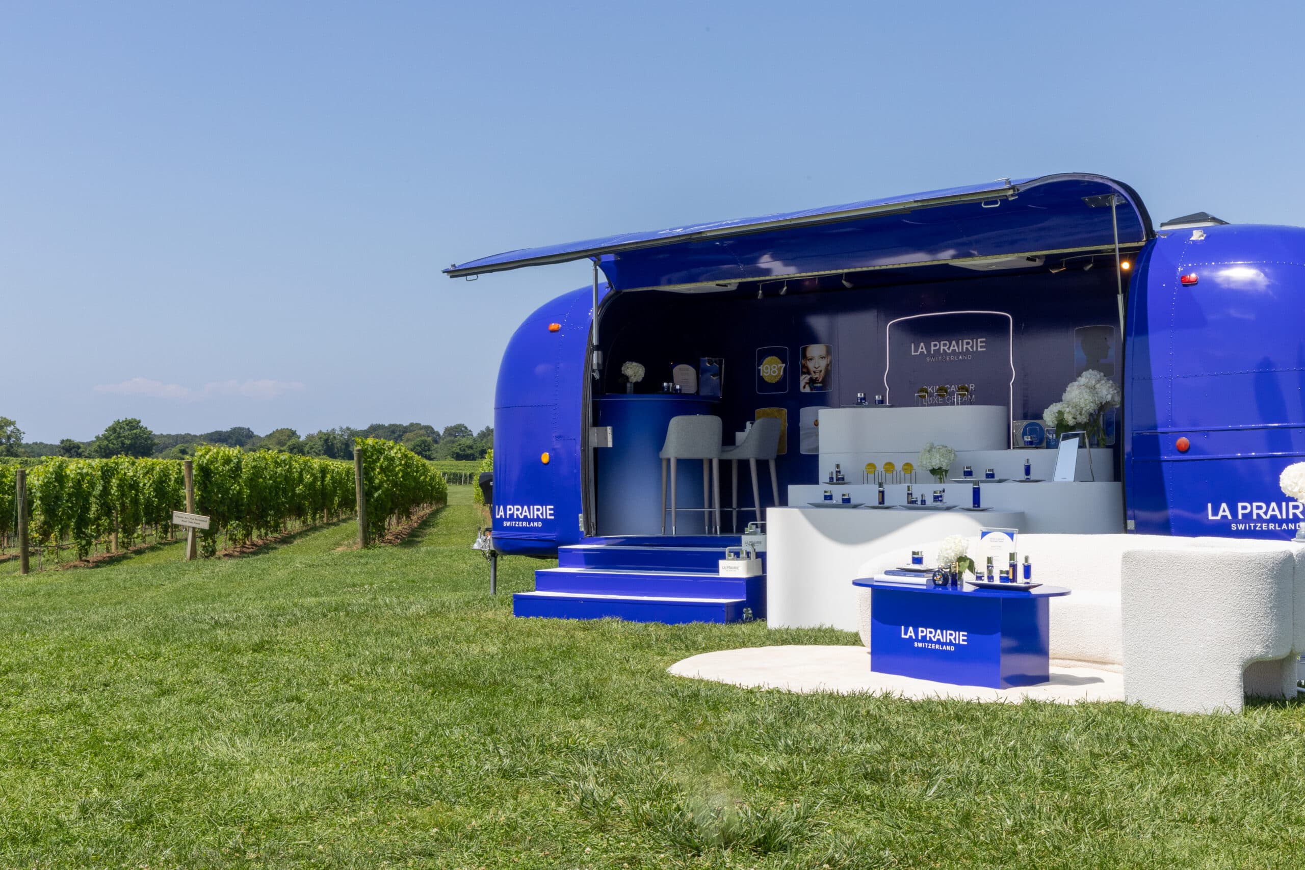 Blue LA PRAIRIE airstream pop-up with skincare products by a vineyard on green grass.