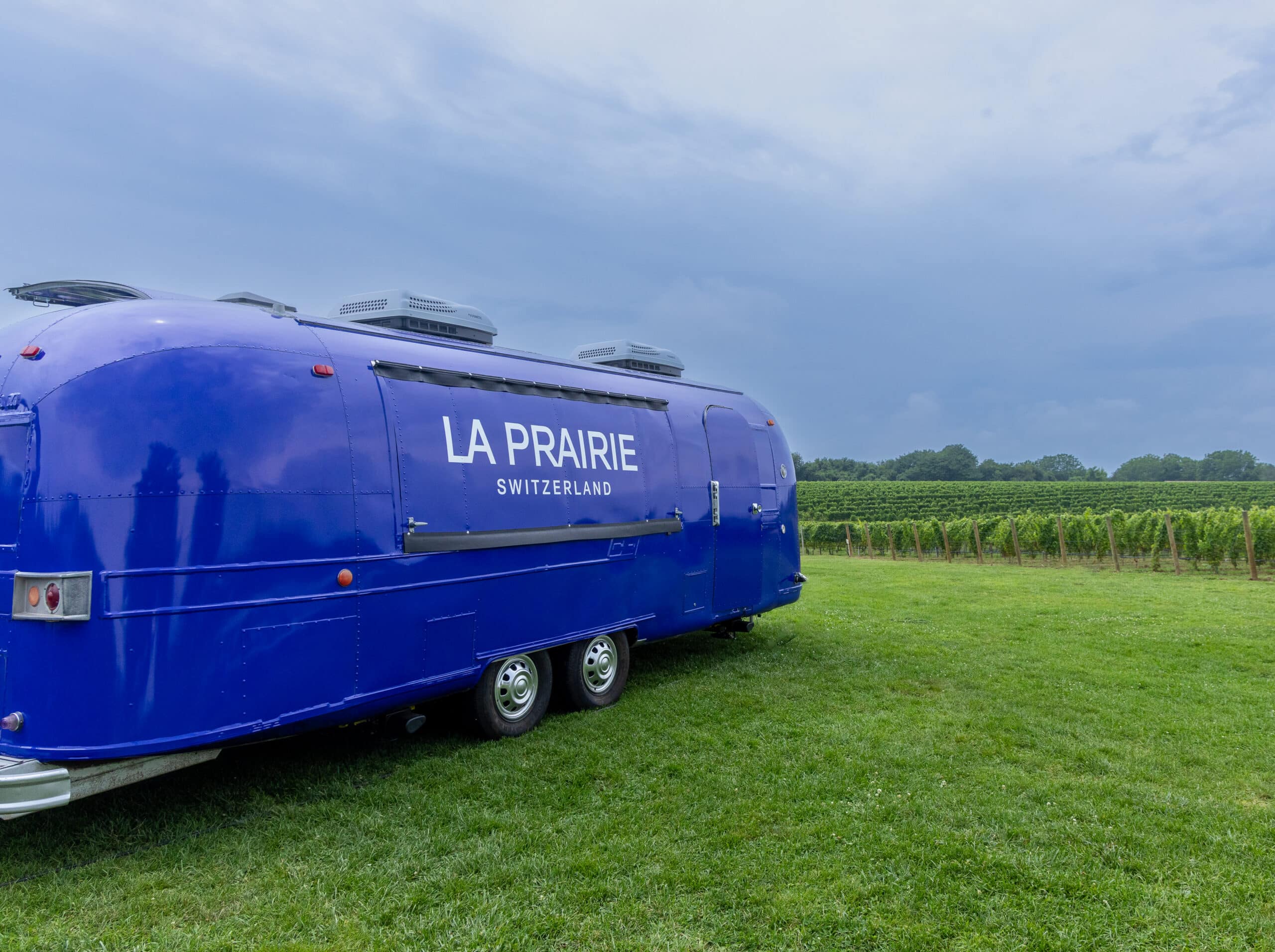 A blue Airstream trailer labeled LA PRAIRIE SWITZERLAND is parked on grass near a vineyard.