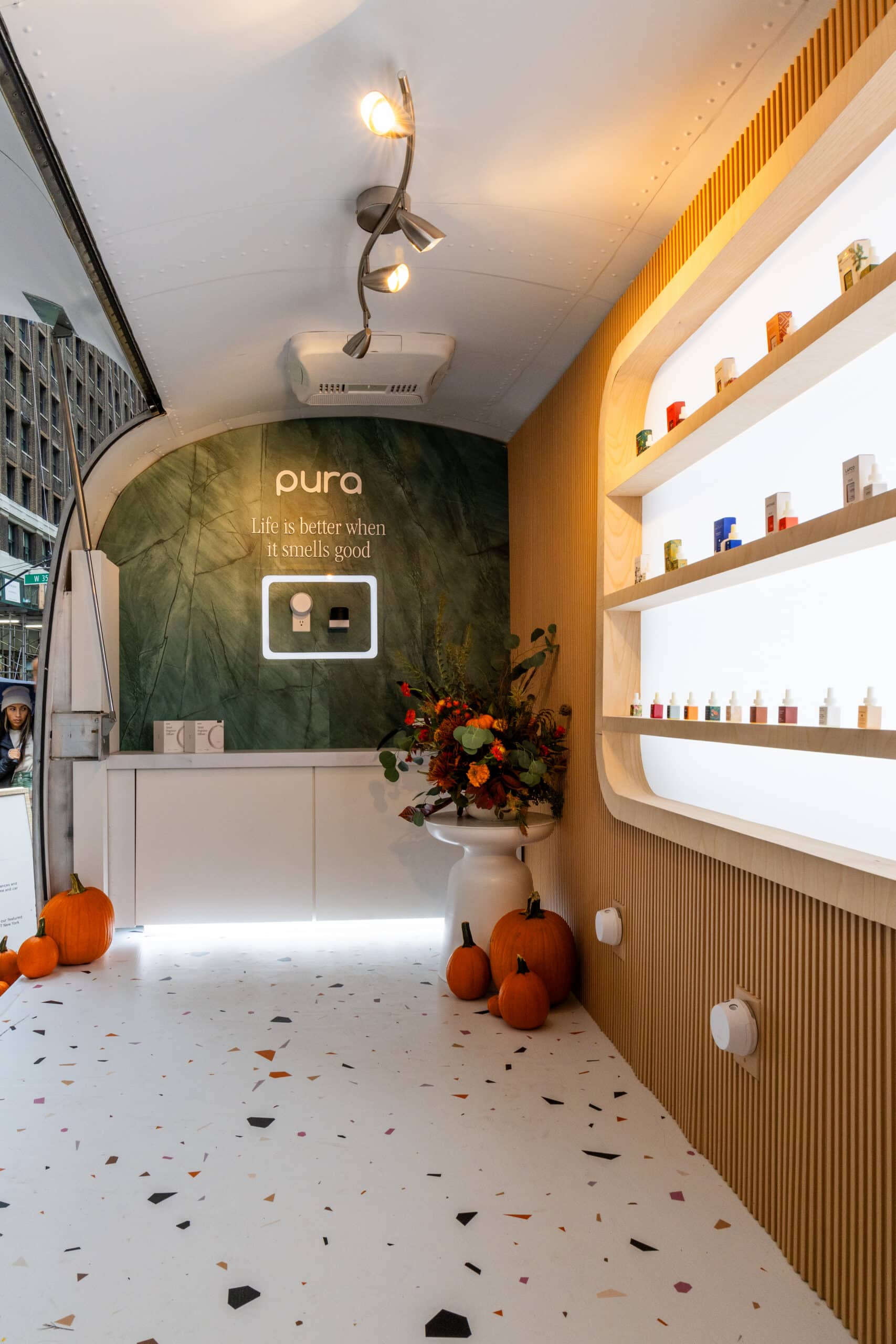 Minimalist room with terrazzo floor, shelves of bottles, flowers, pumpkins, and a green wall sign.