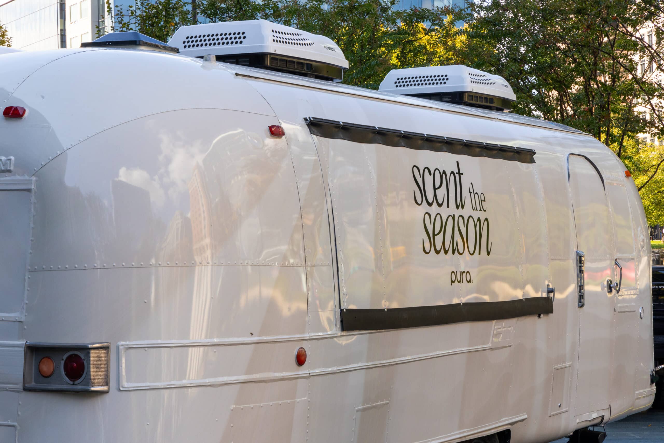 A white airstream trailer with a &ldquo;scent the season pura&rdquo; sign, parked outside with reflections.