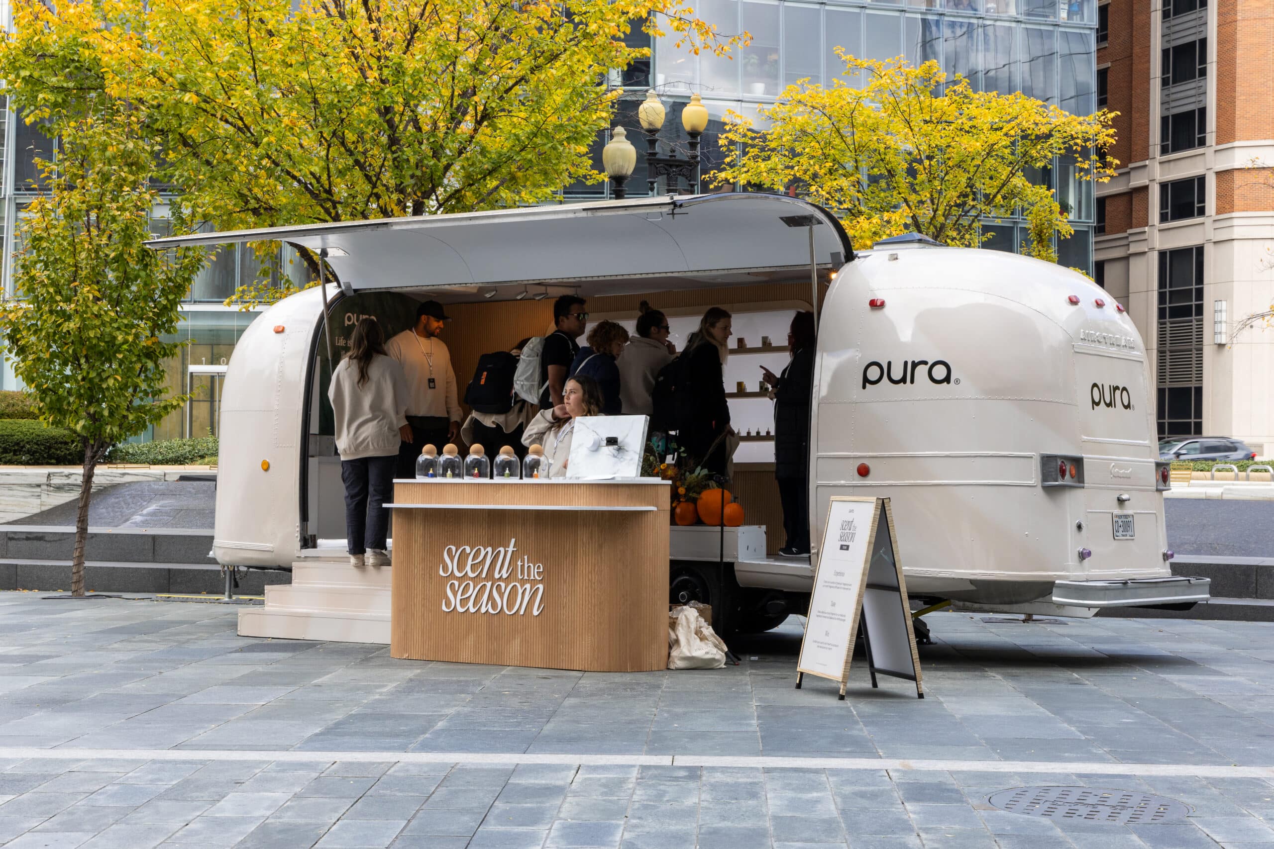 A beige pura. trailer parked on a city street, people inside, scent display and signboard outside.
