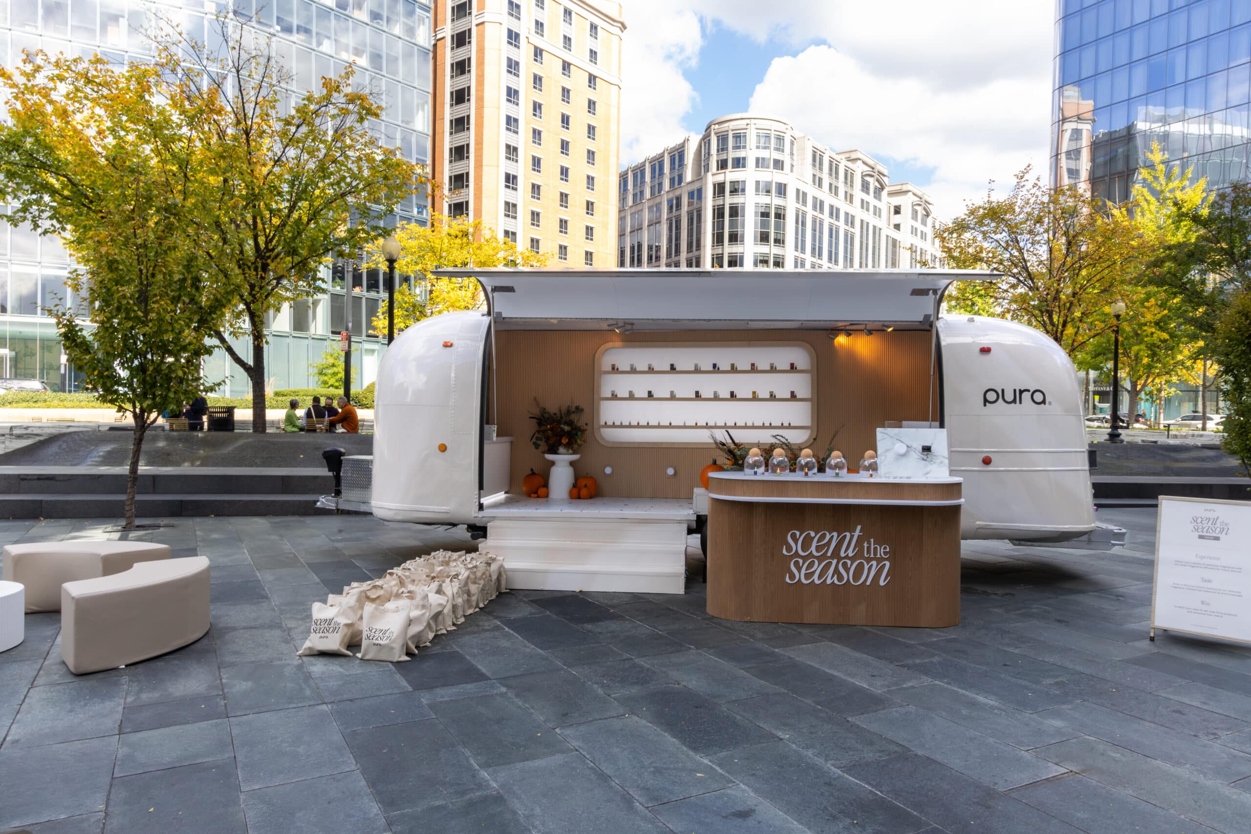 White Pura-branded airstream pop-up with products, gift bags, and fall trees in a city setting.