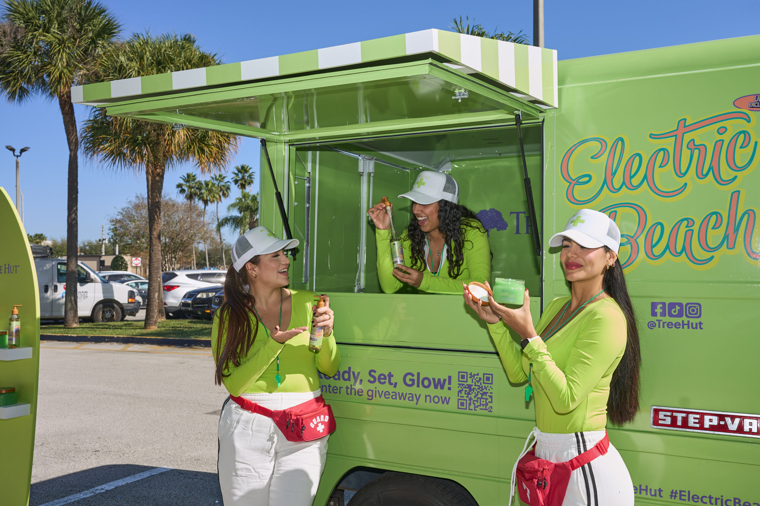 Three women in green tops and white hats eat treats by an "Electric Beach" truck in a sunny lot.