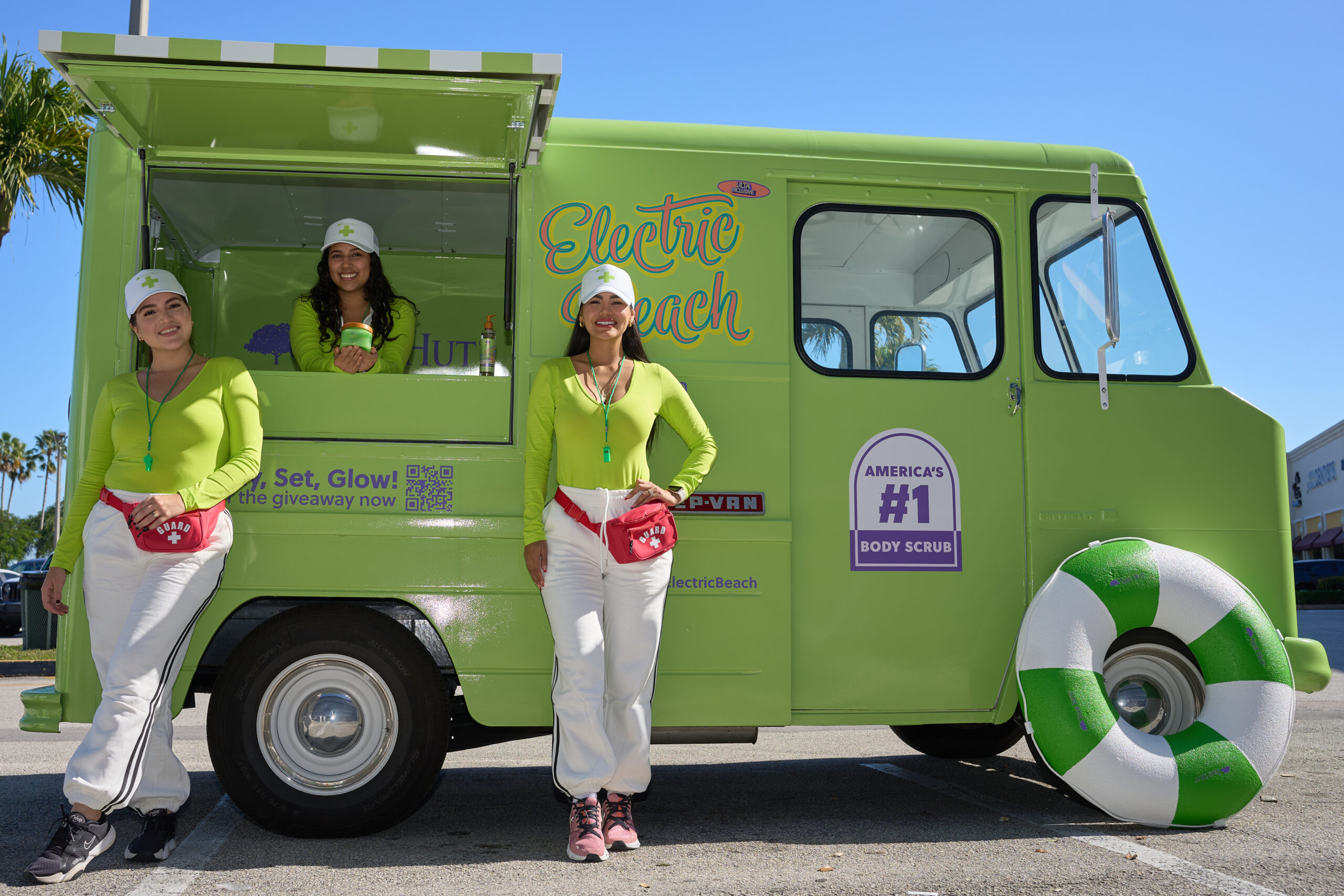 Three women in green shirts and white pants stand by a green Electric Beach truck with signs.