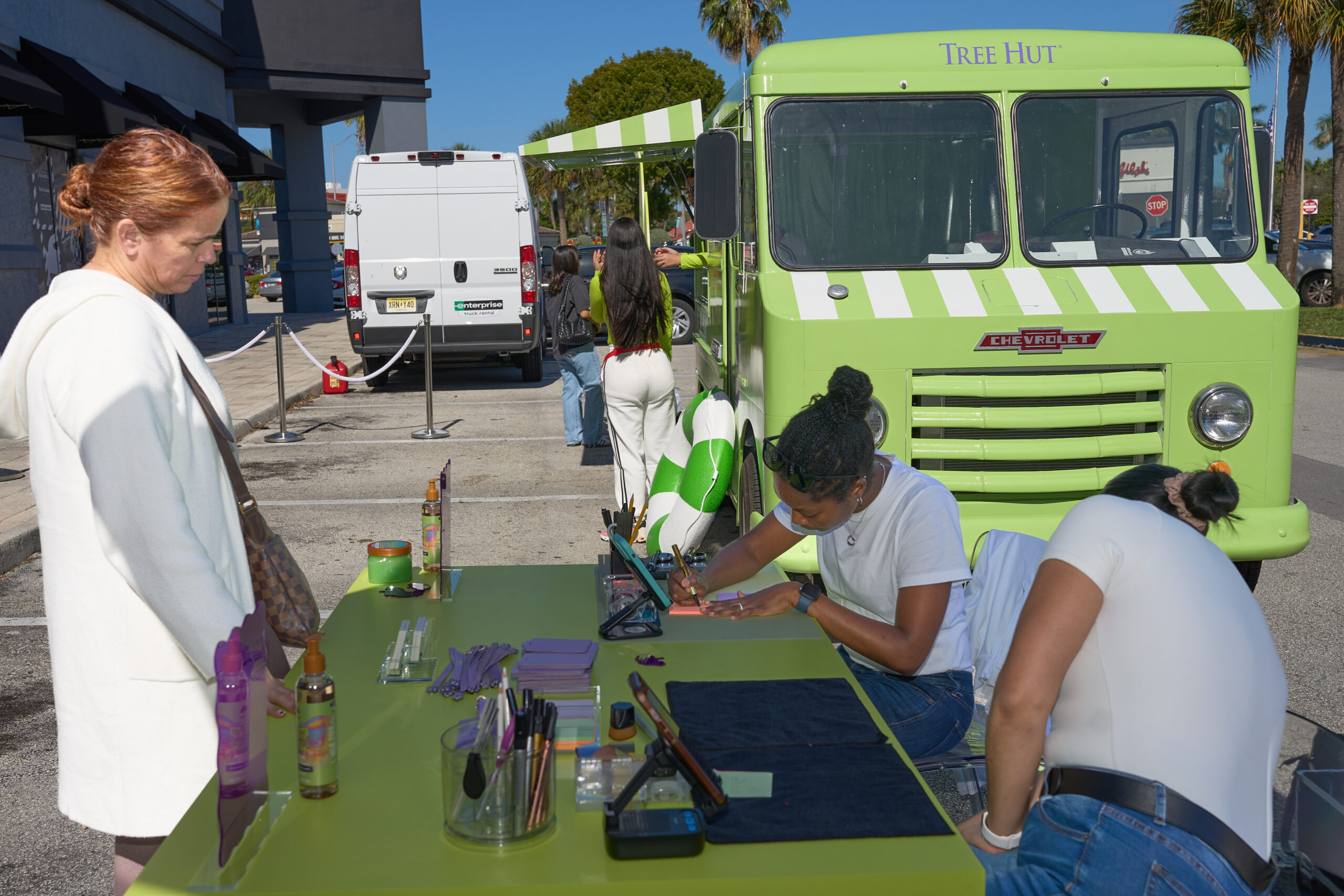 A woman in a white coat watches two women apply makeup at a table near a Tree Hut truck.
