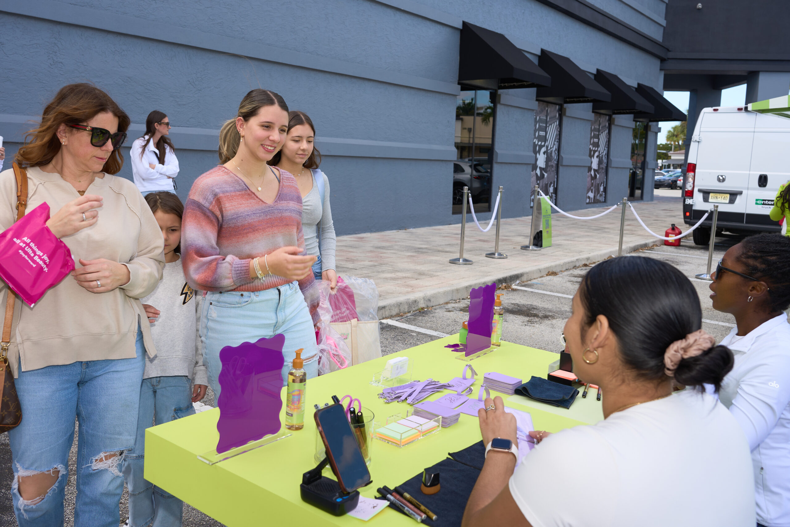 People gather at an outdoor table with display items and promo materials near a gray building.