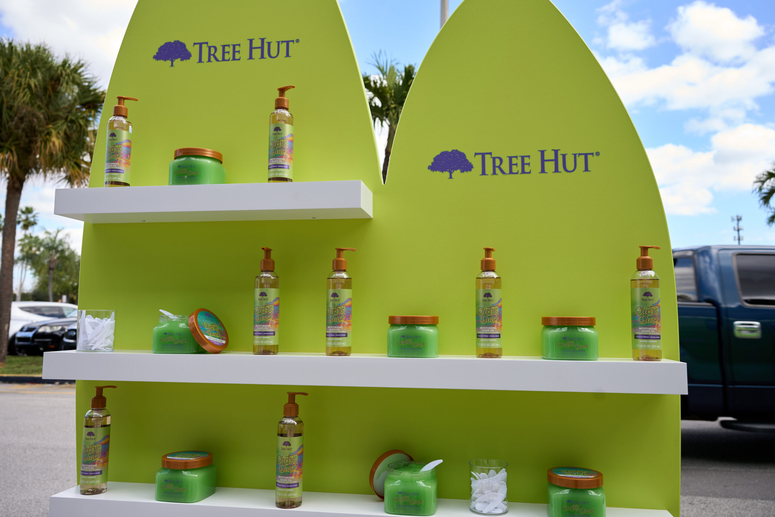 Green display stand with Tree Hut skincare products outdoors, palm trees and blue truck behind.