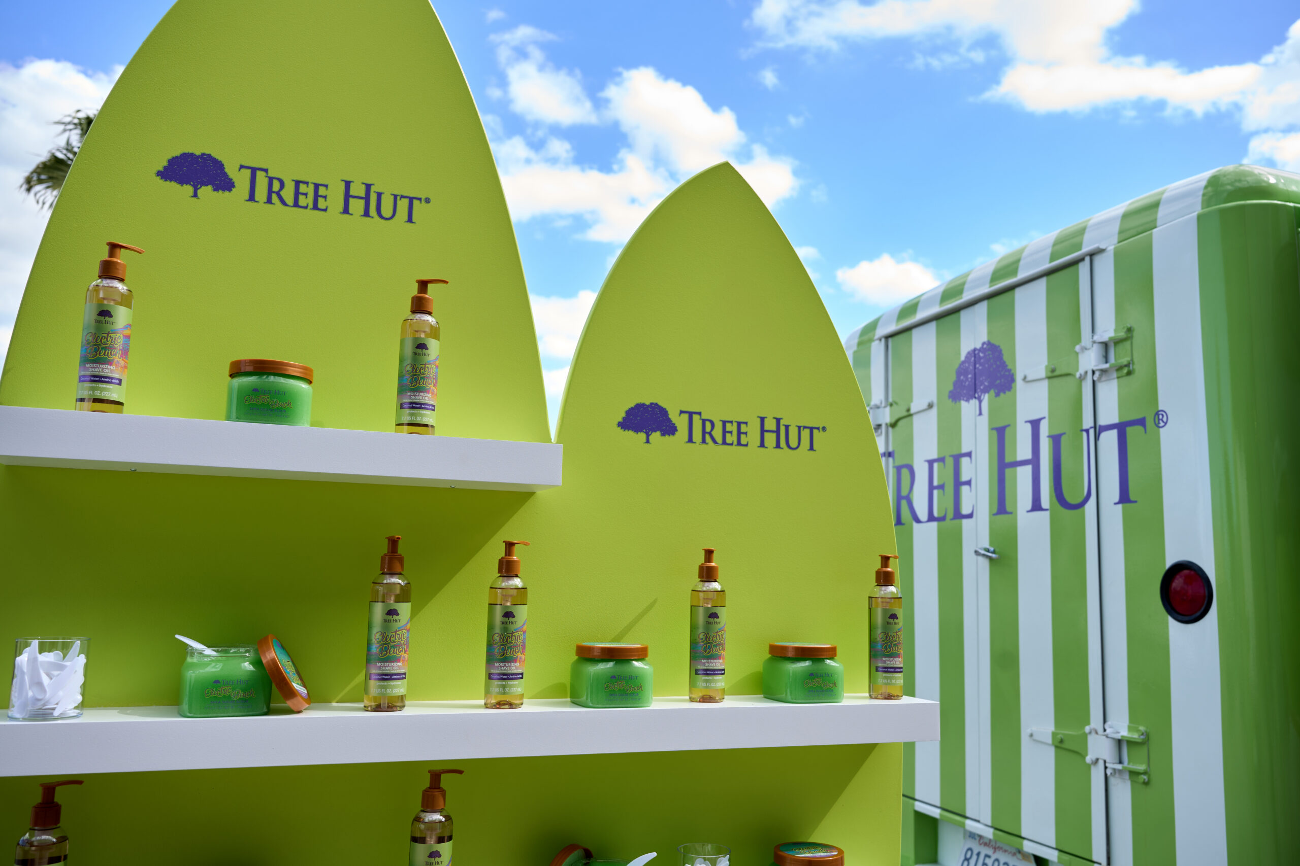 Tree Hut skincare on shelves by green surfboard signs and a striped van under a blue sky.