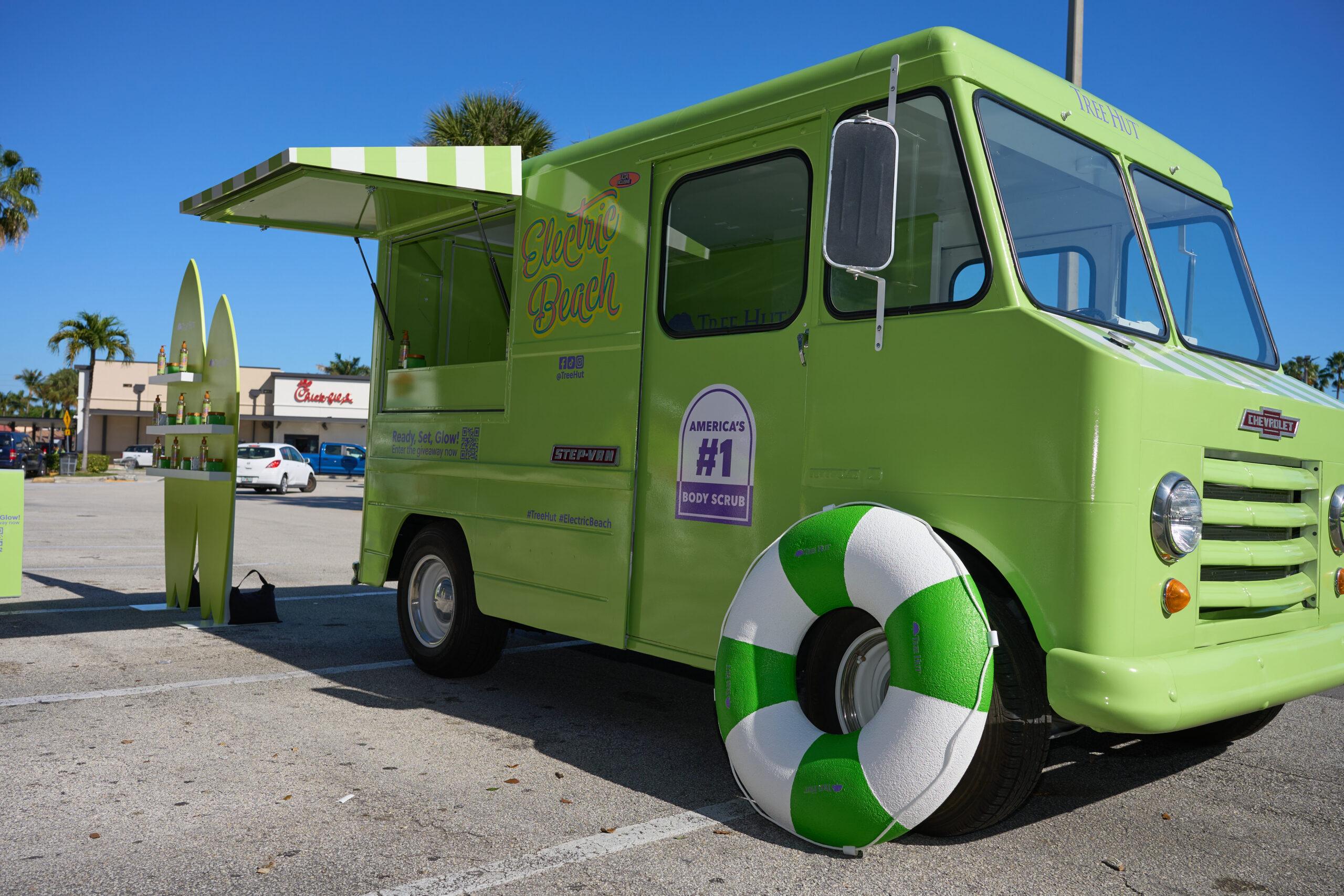 Green food truck with striped awning and lifebuoy, surfboards outside, palm trees behind.