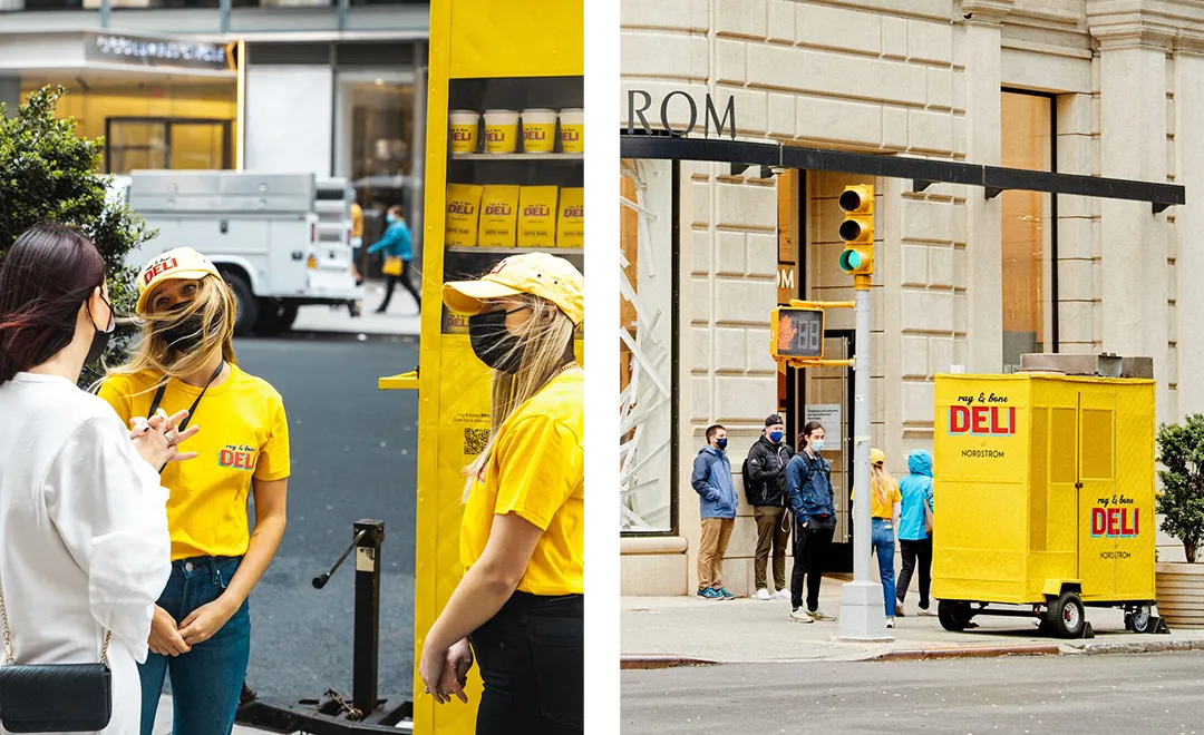 Two women wearing yellow shirts and "New York Deli" caps stand by a bright yellow deli cart on a bustling city street. Engaging with a woman in white, they embody the benefits of experiential marketing as pedestrians and a large building form the backdrop.