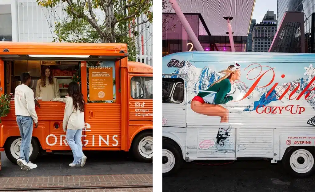 Two adjacent food trucks exemplify experiential marketing: an orange truck branded "Origins," serving two patrons on the left, and a pink-themed truck with a winter design, branded "Pink Cozy Up," on the right, both creating a dynamic scene on a bustling city street.