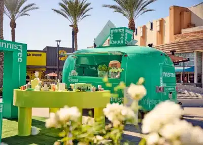 A bright green popup truck with "innisfree" branding embodies the benefits of experiential marketing in an outdoor shopping area. It features a display table with various products. Palm trees and nearby stores, like Buffalo Wild Wings, create a vibrant backdrop.