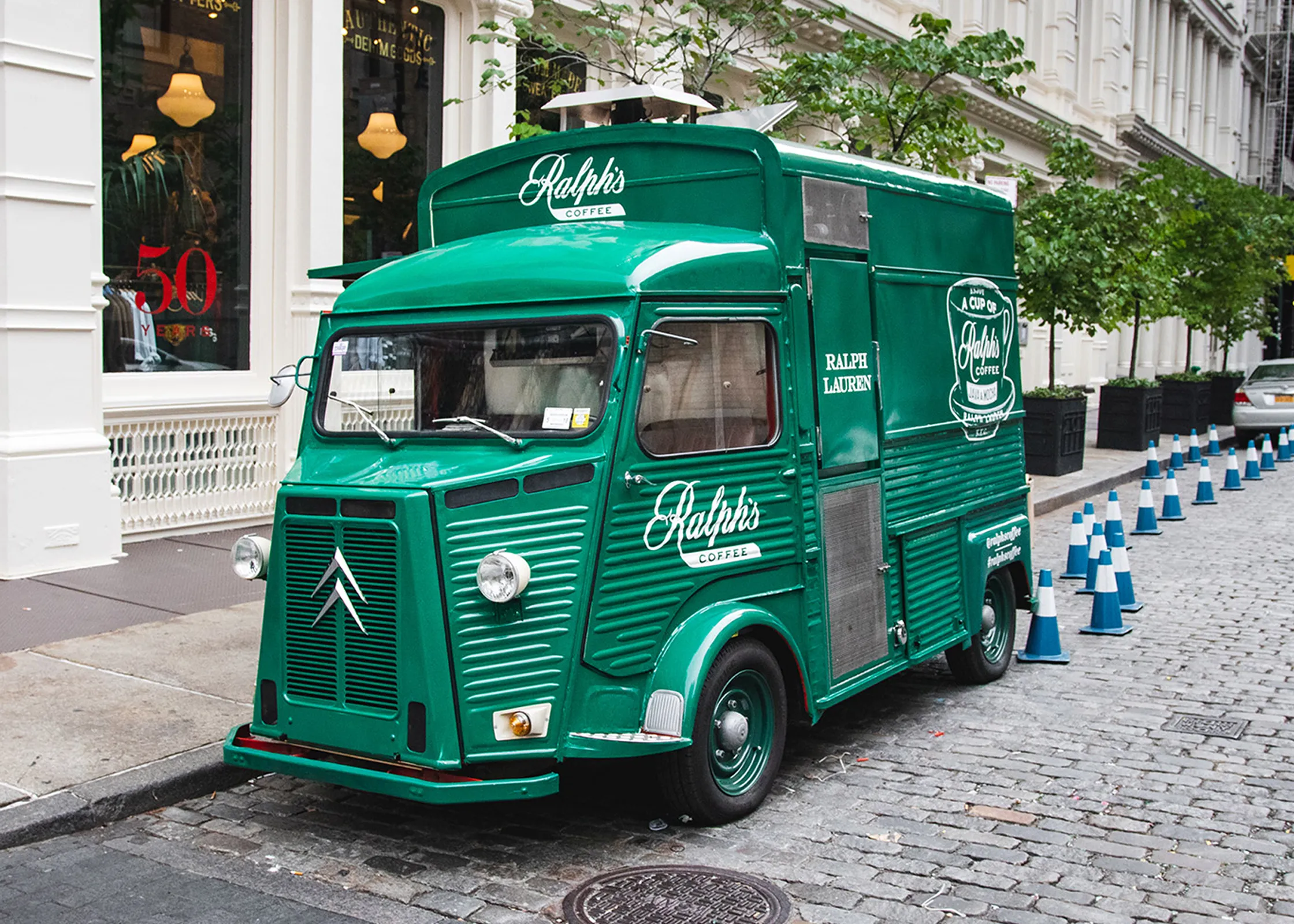 A vintage green food truck adorned with "Ralph's Coffee" and "Ralph Lauren" logos sits on a cobblestone street, exemplifying the creativity of experiential marketing agencies. It's parked near a white building marked by a bold "50," while orange cones neatly line the street.