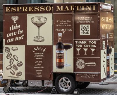 A brown mobile cart on a city street offers espresso martinis. The cart displays graphics of coffee beans, a martini glass, and a bottle, along with promotional text and a QR code. A building with large windows is in the background.