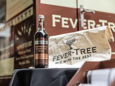 A bottle of Fever-Tree Espresso Martini Mix stands on a black table next to a beige bag with the Fever-Tree logo and the text "Mix With The Best," in front of a brown and white Fever-Tree branded backdrop.