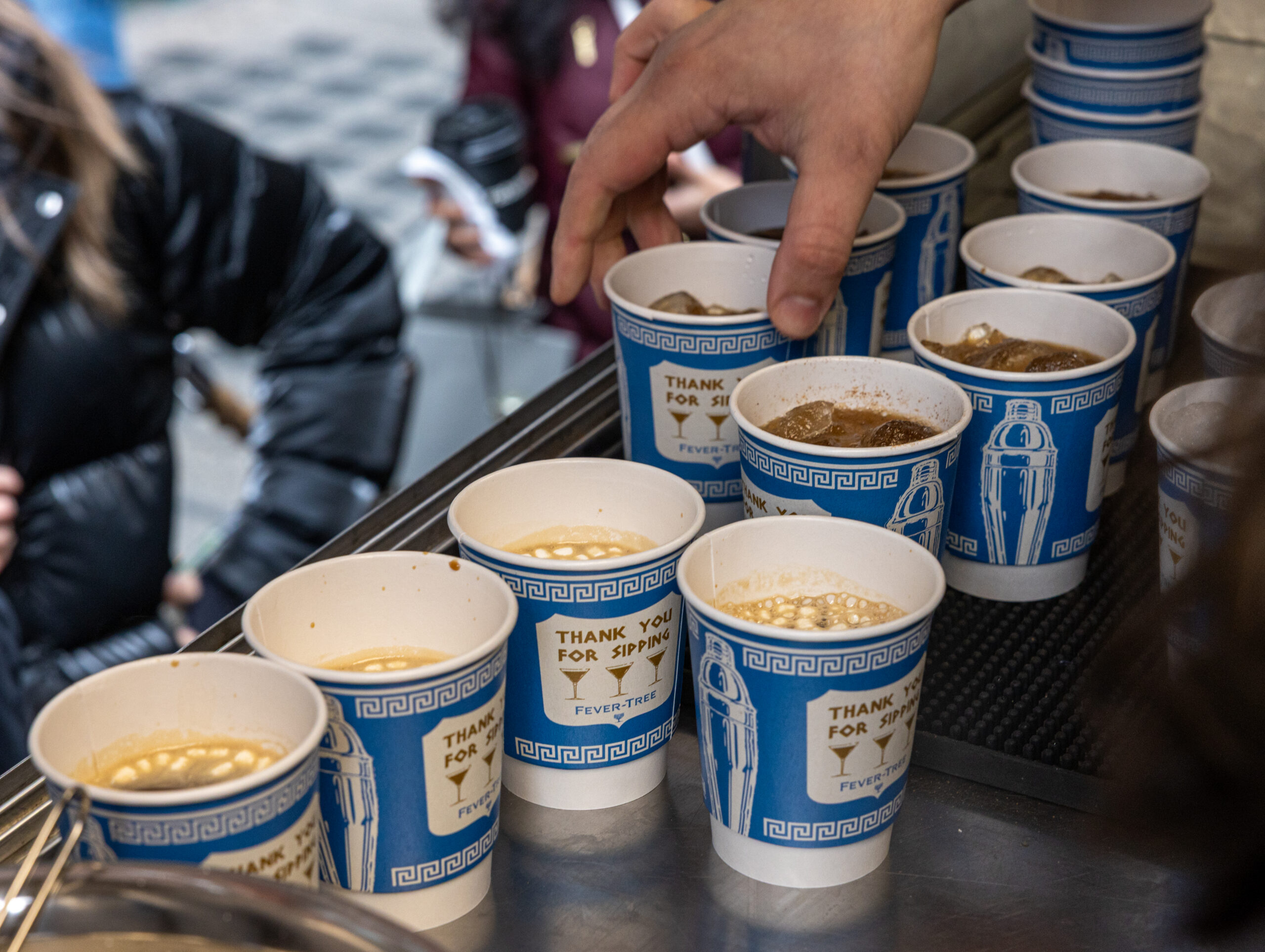 A hand reaches for a blue and white coffee cup on a counter; people are blurred behind.