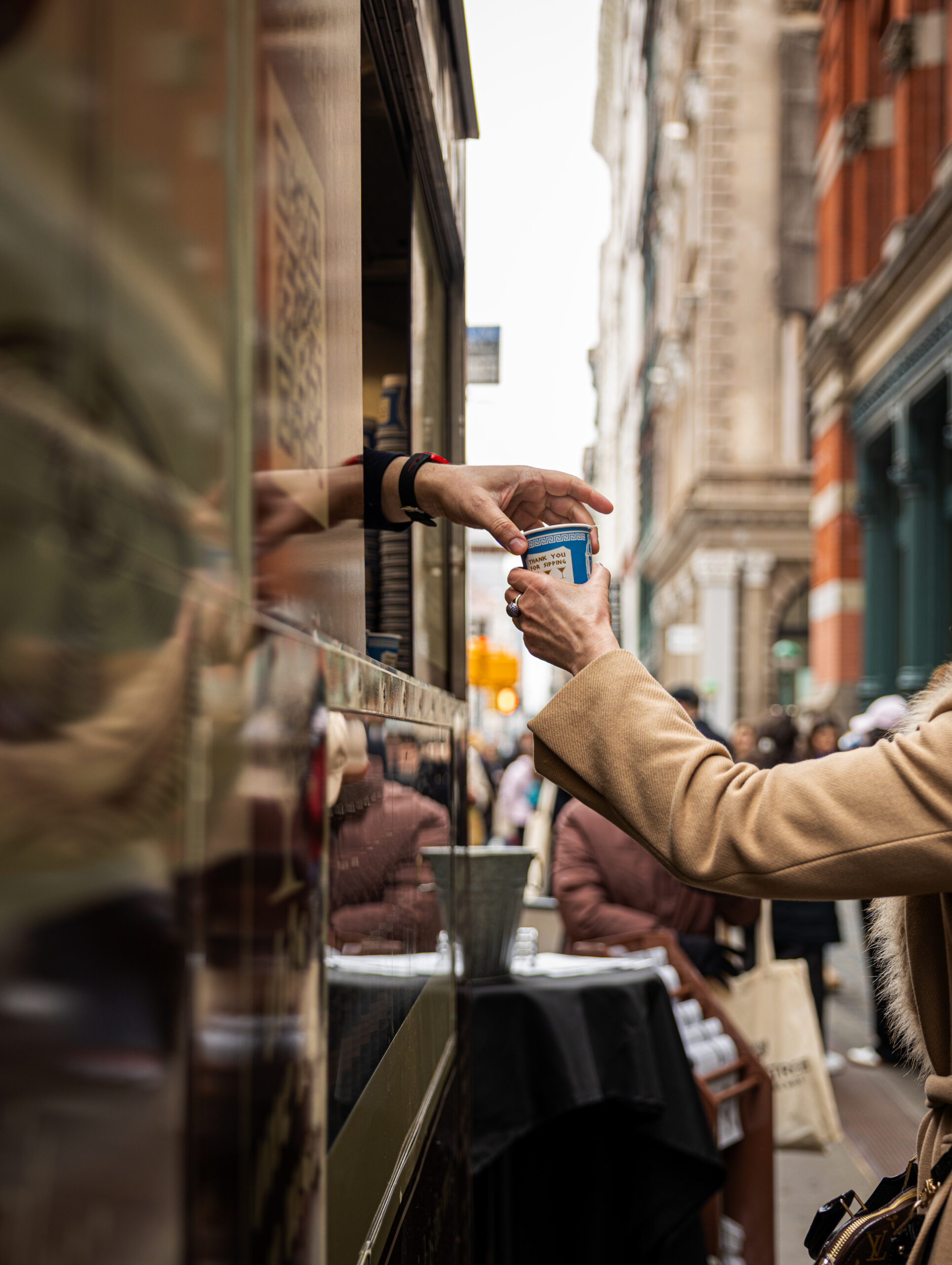 Person in tan coat takes coffee from vendor at food truck, city street and old buildings behind.