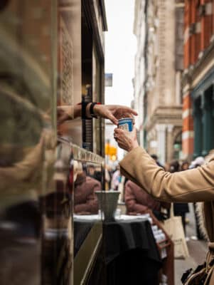 A person in a tan coat receives a cup of coffee from a server through a food truck window on a busy city street. The background shows blurred buildings and pedestrians.