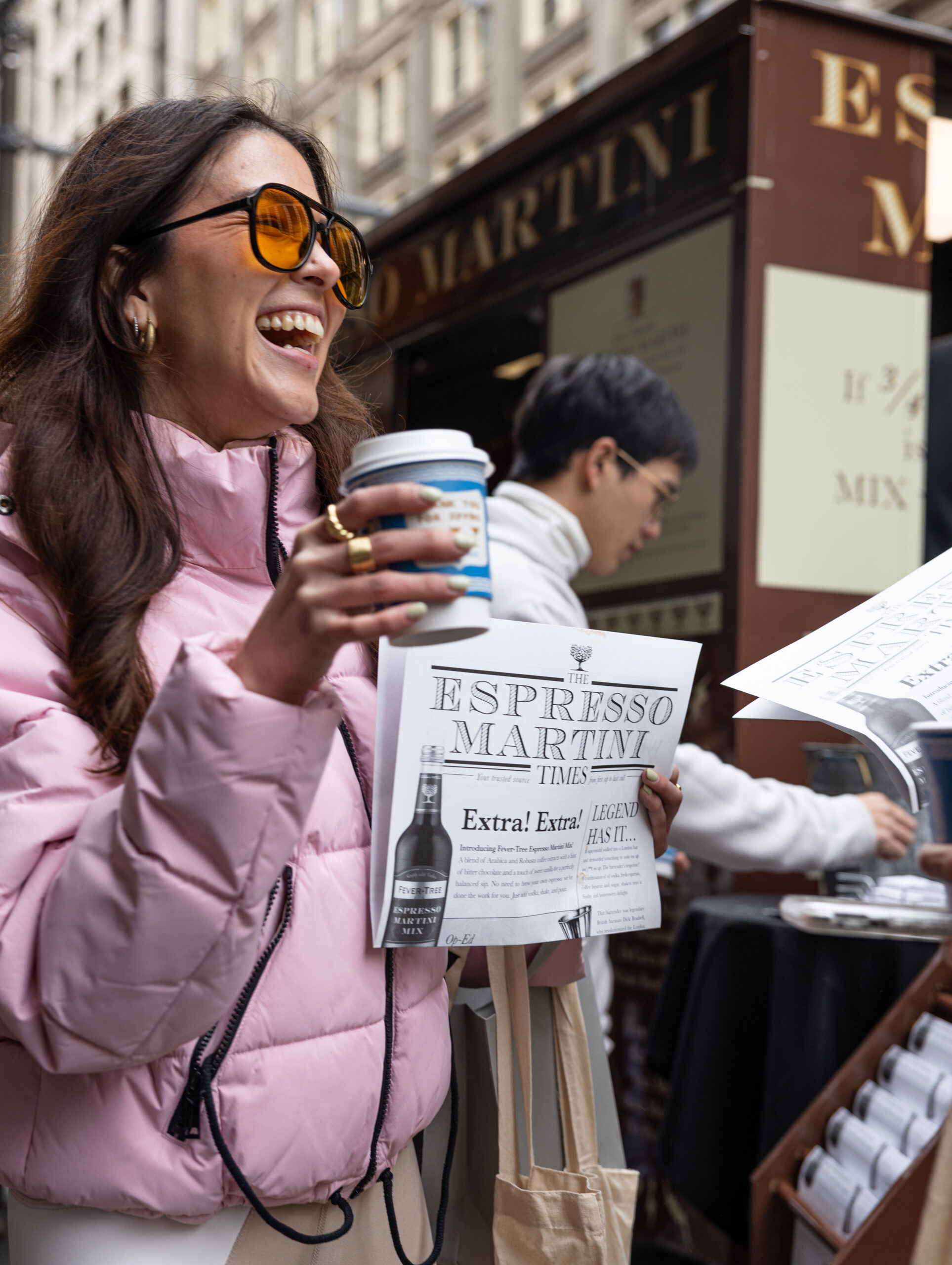 Woman in pink jacket with sunglasses holds coffee and newspaper on city street; kiosk nearby.