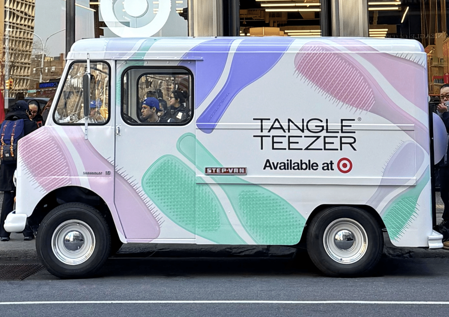 Tangle Teezer A pastel-colored van with "Tangle Teezer Available at Target" parked in front of a Target store on a city street. People are visible through the store windows in the background.
