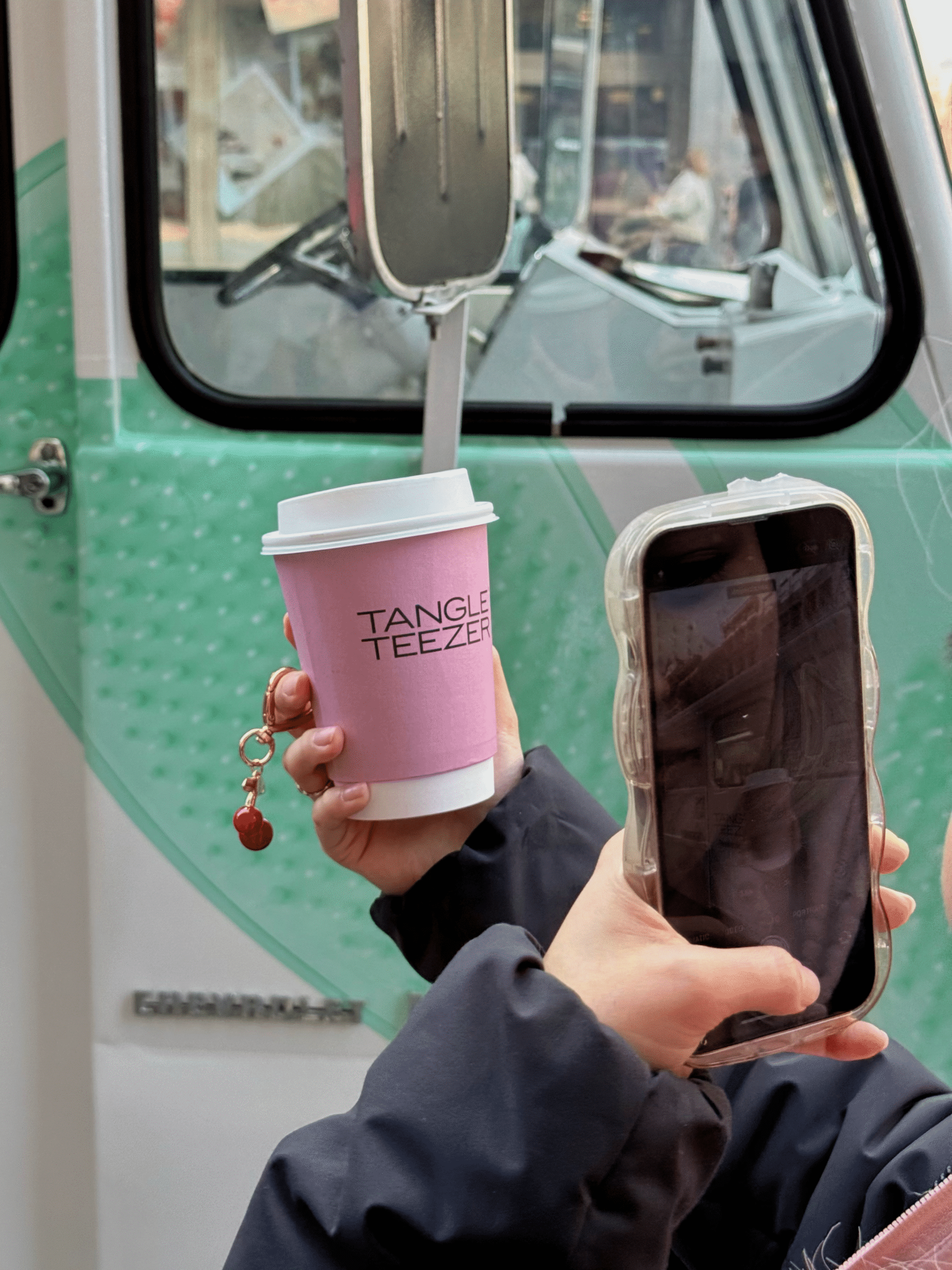 Person holding pink Tangle Teezer cup and smartphone stands before mint green and white vehicle.