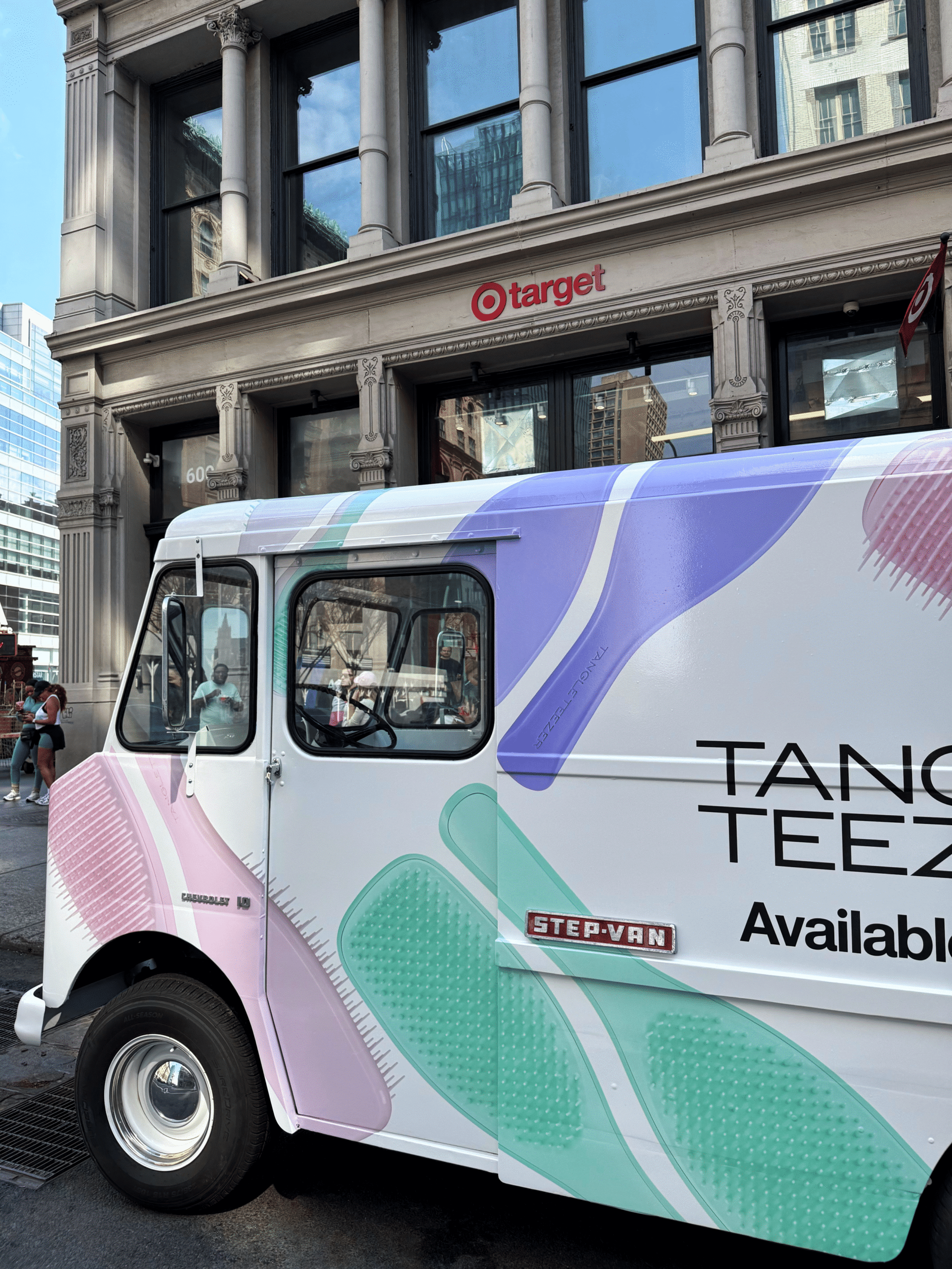 Pastel van marked “Tangle Teezer Available” parked outside Target on a city street.