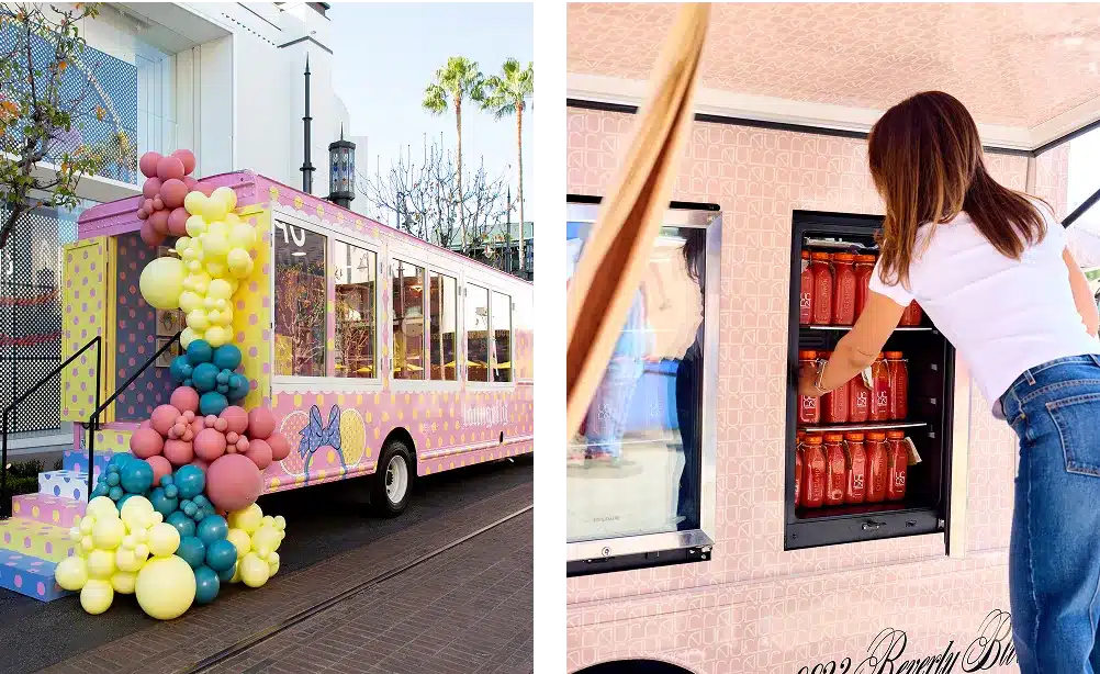 A pink and yellow polka dot bus decorated with colorful balloons is parked on a street. A woman reaches into a vending window on the bus to grab a red drink bottle.