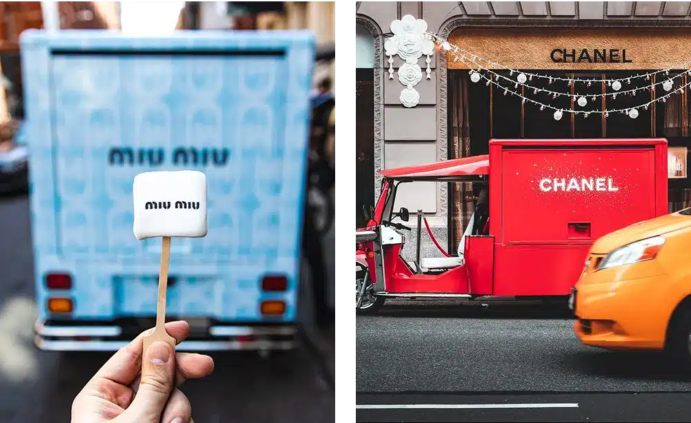 A hand holds a marshmallow with "miu miu" printed on it in front of a blue Miu Miu truck; beside it is a red Chanel vehicle parked on a street near a Chanel store, with a yellow taxi passing by.