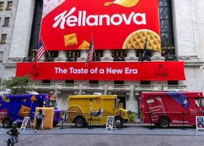 Three colorful Experiential Marketing Vehicles from Kellanova are parked outside the New York Stock Exchange, with large banners displaying the Kellanova logo and the slogan “The Taste of a New Era” above them. Two American flags are visible.