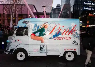 A vintage truck painted with a winter mountain scene and a woman in ski gear serves as an Experiential Marketing Vehicle for "Pink Cozy Up" by Victoria's Secret Pink, parked on a busy city street surrounded by people and tall buildings.