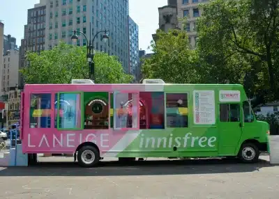 A colorful beauty product truck, part of Experiential Marketing Vehicles, with “LANEIGE” in white on a pink section and “innisfree” on green, is parked in a city plaza surrounded by trees and tall buildings.