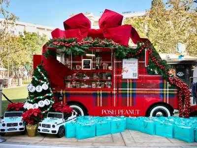 A festive red food truck, showcasing experiential marketing strategies, is decorated with garland, ornaments, and a giant red bow. “POSH PEANUT” shines on the side, surrounded by turquoise gift bags and two white toy cars at an outdoor event.
