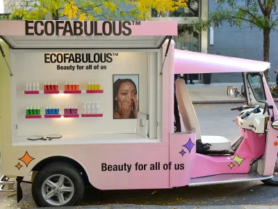 A pink three-wheeled beauty truck displays colorful skincare products and a poster of a woman touching her face. The words "ECOFABULOUS" and "Beauty for all of us" are printed on the truck’s side.
