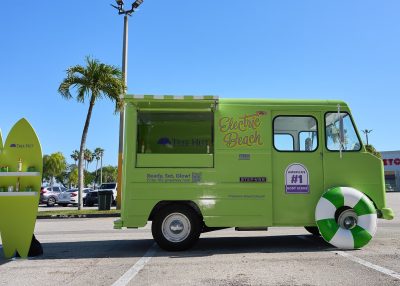 A bright green food truck with "Electric Beach" branding is parked outside under palm trees, perfect for mobile marketing tours. A matching green surfboard display with products stands beside it, and the truck sports a beach ball hubcap on the front wheel.
