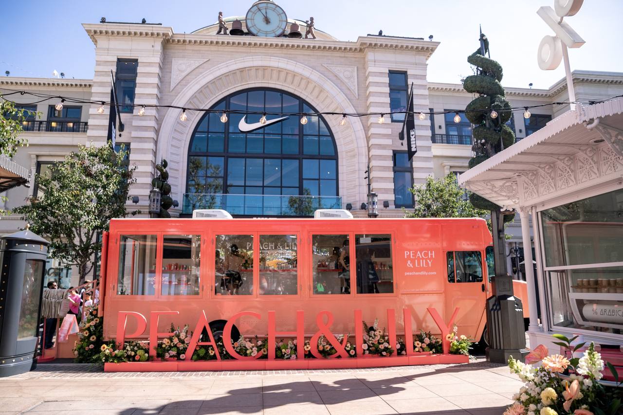 A bright peach-colored PEACH & LILY pop-up booth, created by an Experiential Marketing agency, is set up in front of a grand Nike building, surrounded by flowers and outdoor string lights under a clear blue sky.
