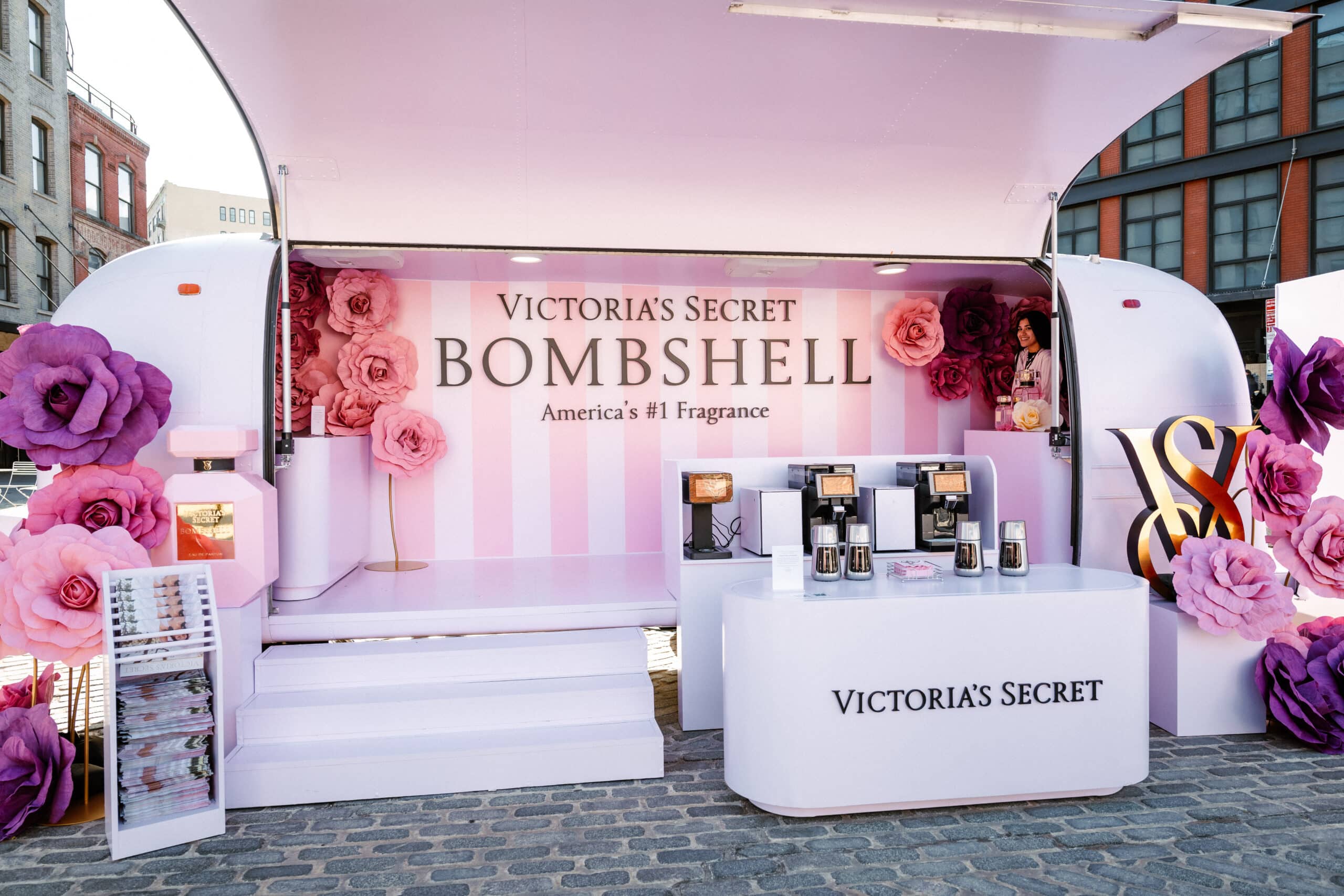 Victoria’s Secret Bombshell pop-up with pink flowers and signs on a cobblestone street outdoors.