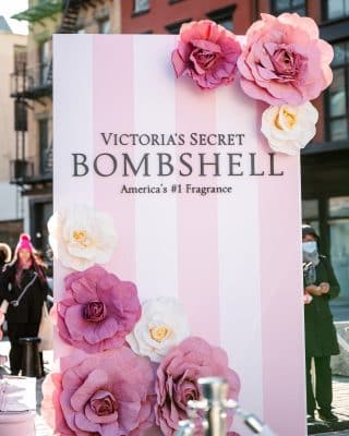 A large pink and white striped display sign reads "Victoria’s Secret Bombshell, America’s #1 Fragrance," decorated with oversized paper flowers in pink, purple, and white. People stand in the background outdoors.
