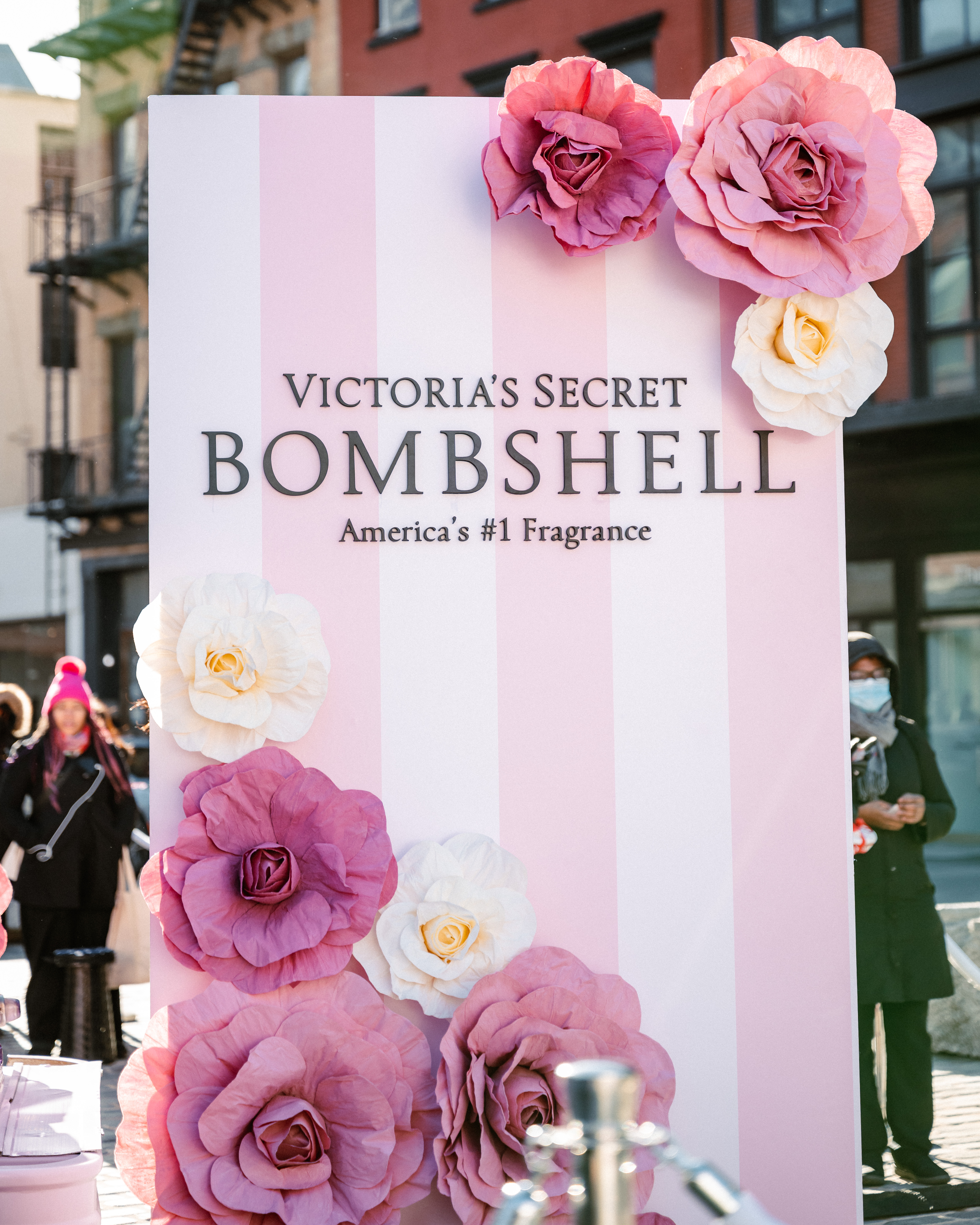 A large pink and white striped display sign reads "Victoria’s Secret Bombshell, America’s #1 Fragrance," decorated with oversized paper flowers in pink, purple, and white. People stand in the background outdoors.
