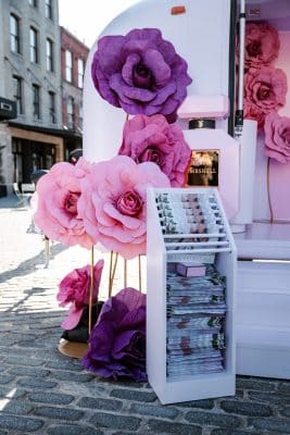 A display of large, decorative pink and purple paper flowers stands next to a white magazine rack filled with publications on a cobblestone street, with buildings visible in the background.