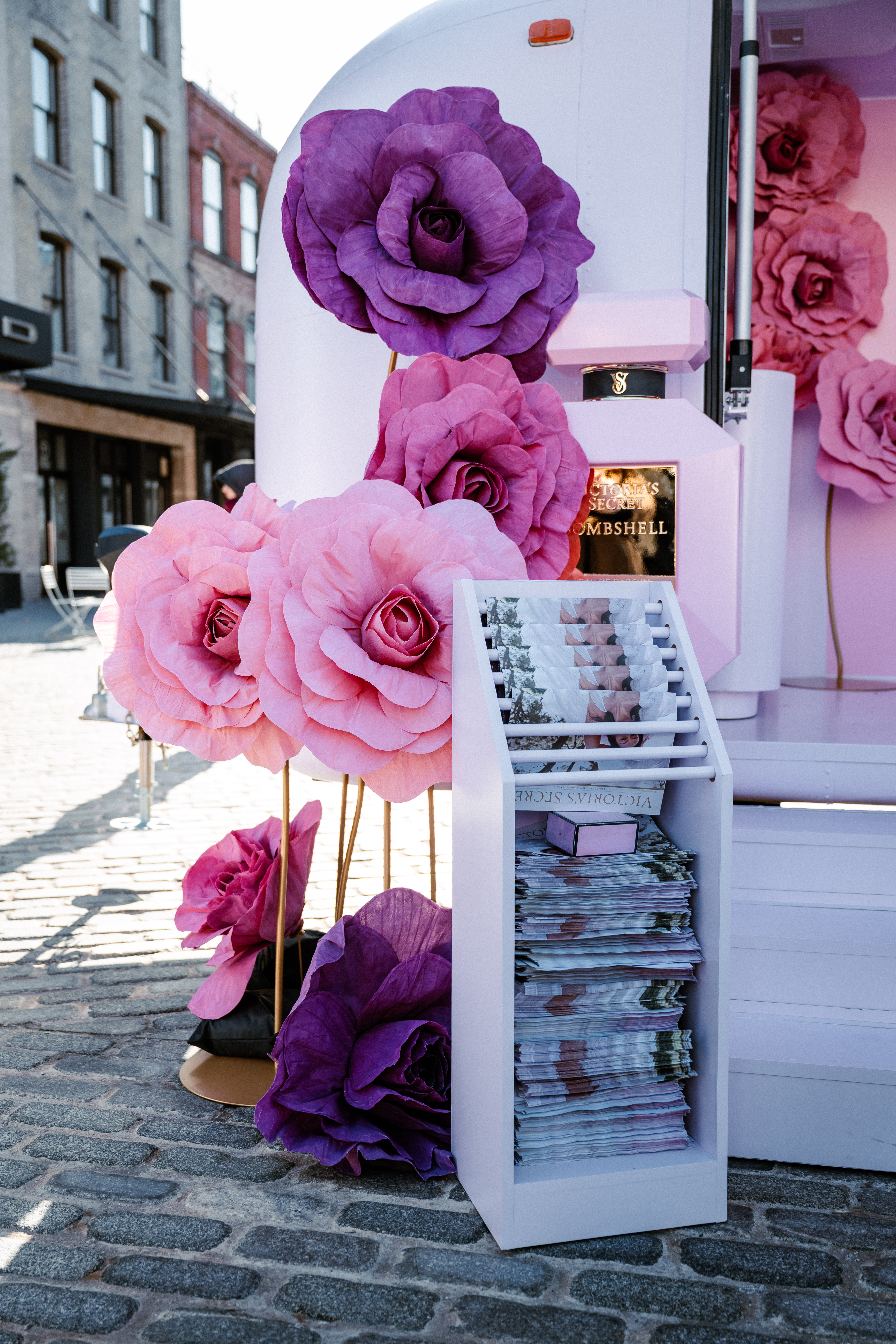 A display of large, decorative pink and purple paper flowers stands next to a white magazine rack filled with publications on a cobblestone street, with buildings visible in the background.